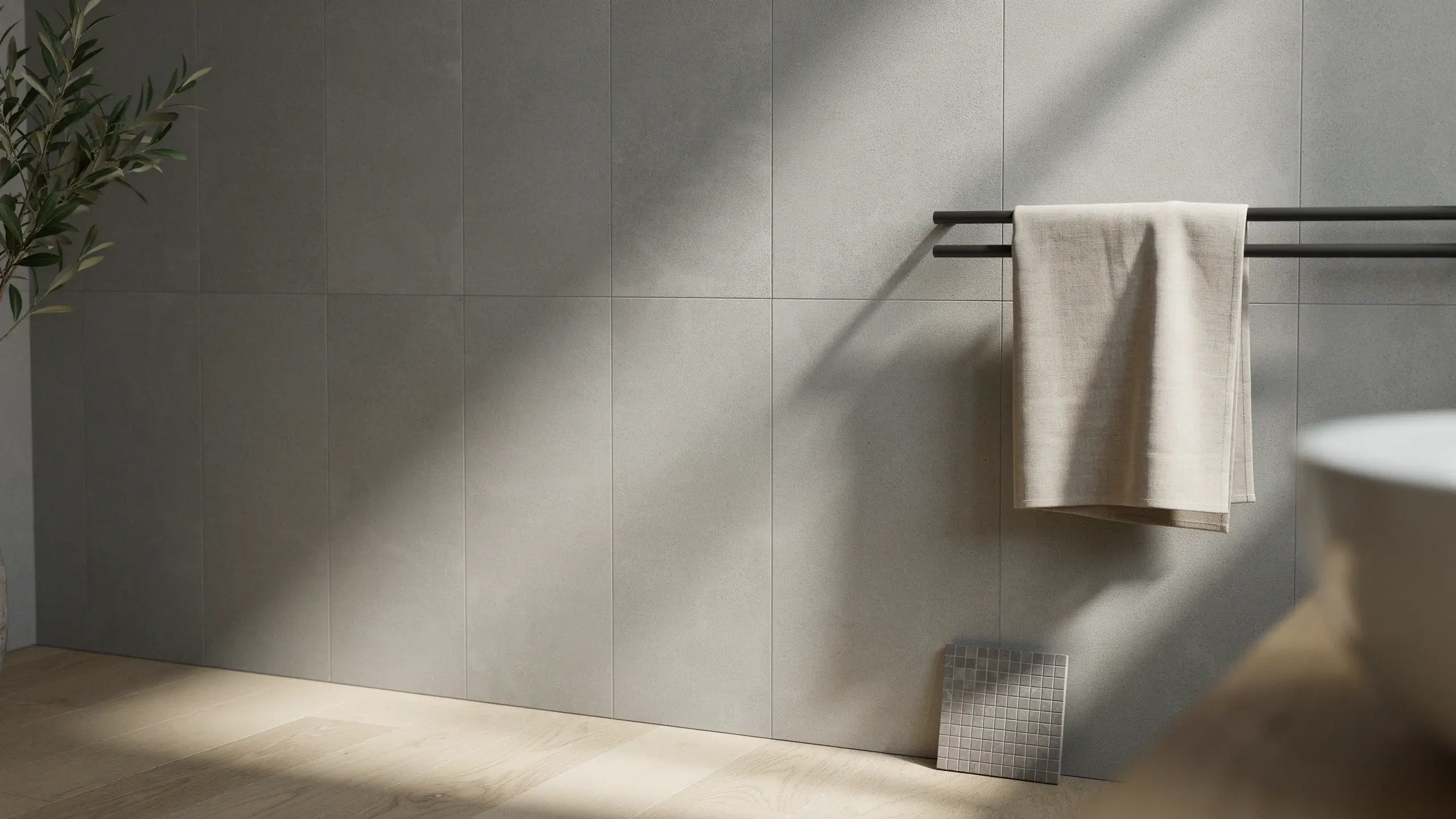 Modern minimalist bathroom interior featuring expansive light-grey matte wall tiles, contrasting with a small square mosaic tile sample displayed on the wooden floor next to a black towel rail.