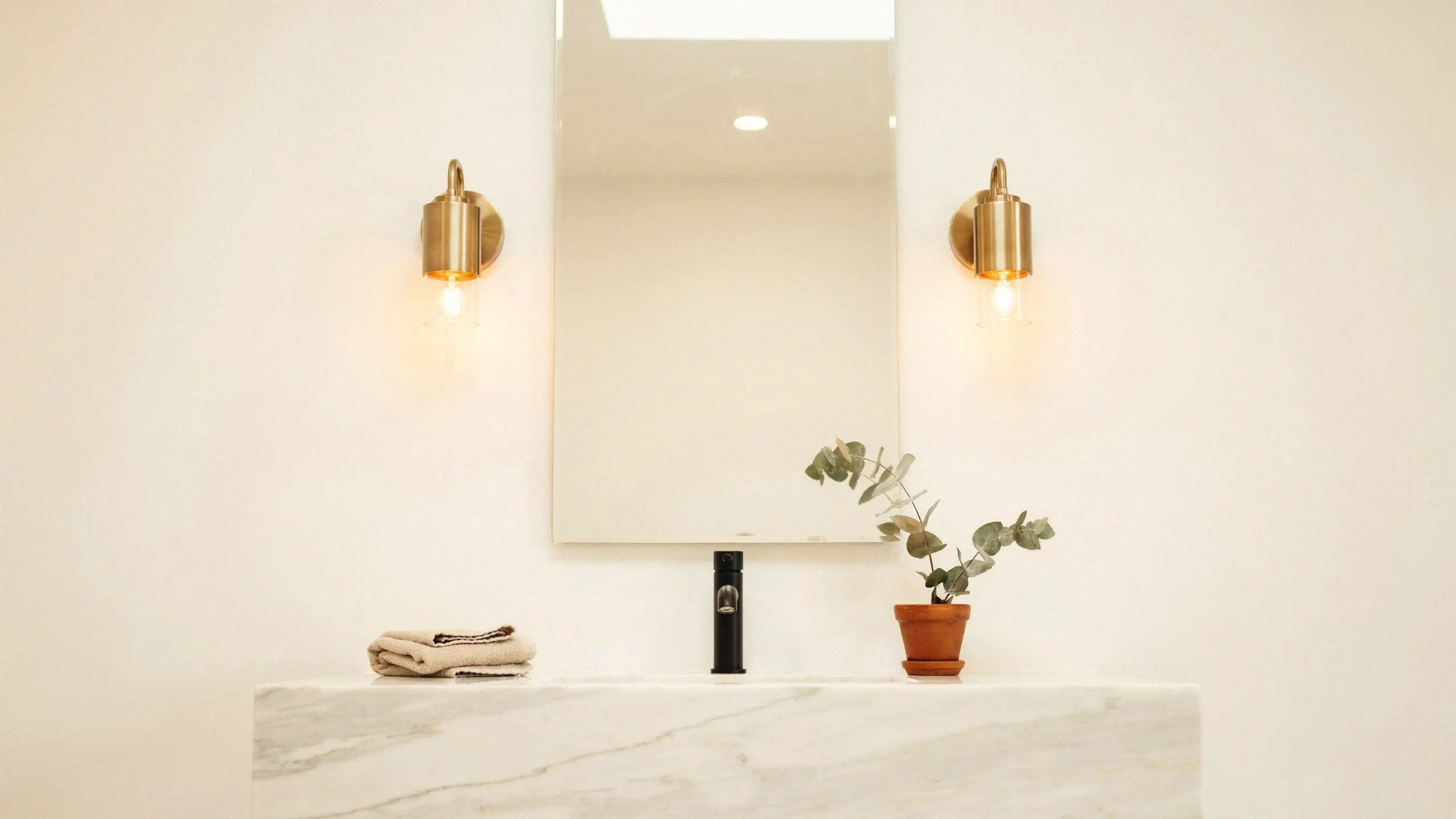Warm brass wall sconces providing layered lighting above a minimalist marble vanity with a black faucet and potted eucalyptus, illustrating how to replace a single ceiling bulb.