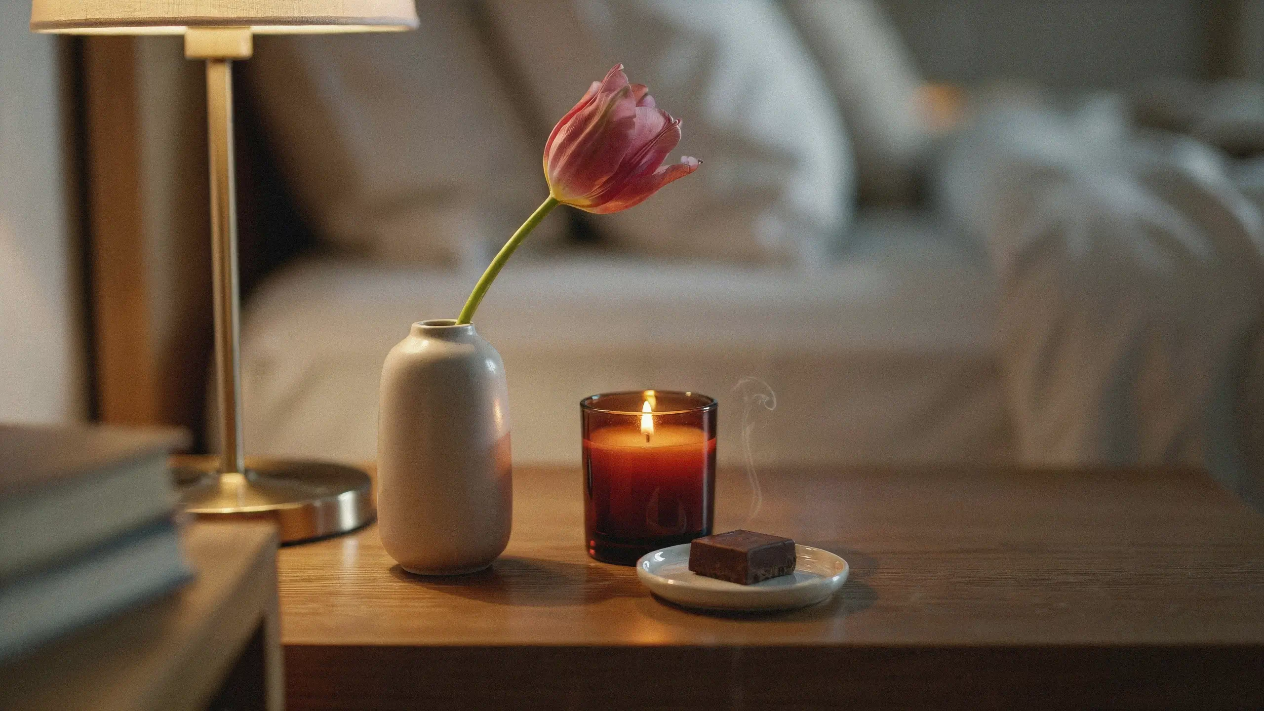 Stylish bedside tray with a pink tulip, glowing amber candle, and dark chocolate, styled like a luxury hotel nightstand.