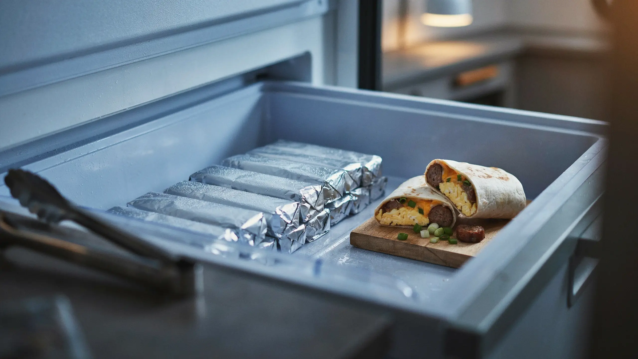 Frozen breakfast burritos wrapped in aluminum foil stacked in an open freezer drawer, with one burrito cut open on a small wooden board showing fluffy scrambled eggs, sausage, melted cheddar cheese, and green onion garnish.