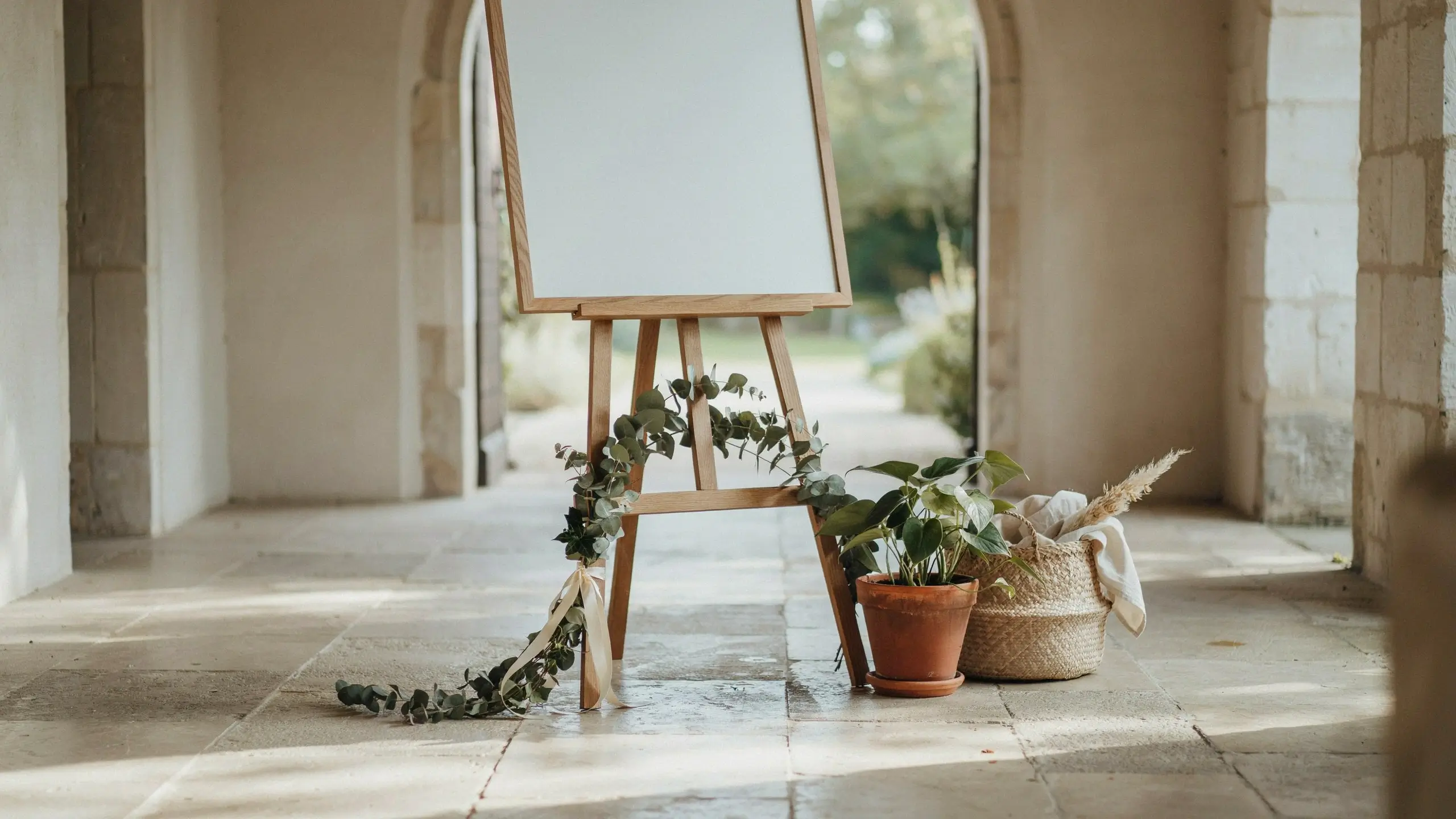 A wooden MÅLA easel holding a blank white sign board used as a wedding welcome sign or seating chart, elegantly decorated with a draping eucalyptus garland and cream ribbon, positioned next to a terracotta potted plant and a woven basket with pampas grass in a sunlit stone archway.