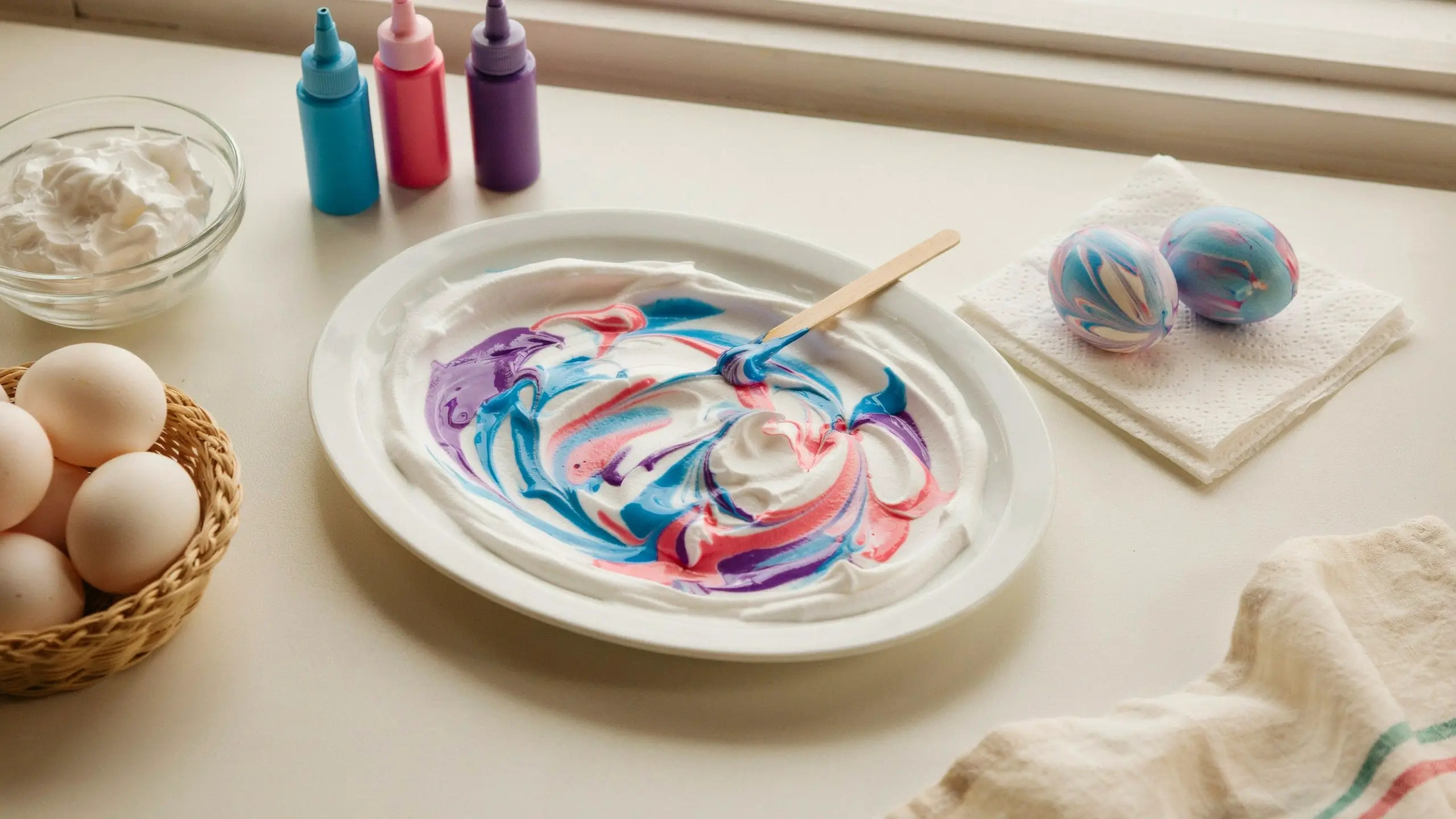 A bright DIY setup for making marbled Easter eggs using shaving cream and food coloring, featuring swirled cream, dye bottles, a basket of white eggs, and two finished marbled eggs on a paper towel.