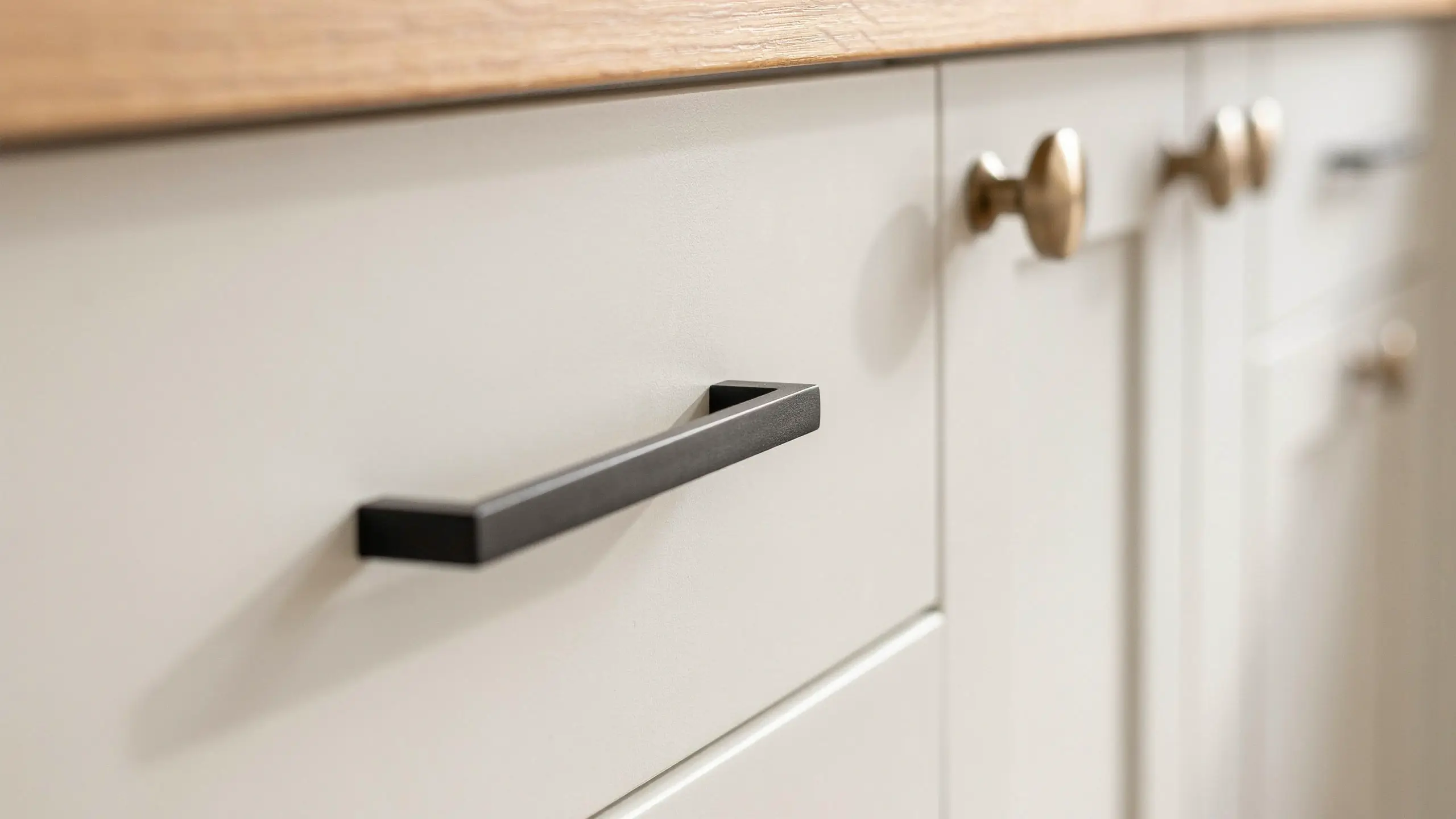 Close-up of a modern matte black rectangular handle and brass round knobs on white shaker kitchen cabinets, illustrating an affordable hardware upgrade.