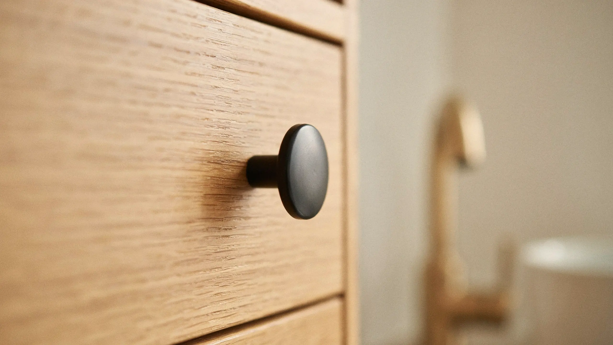 A close-up of a matte black, round drawer knob mounted on a light oak wood cabinet drawer front, demonstrating the use of muted and understated metal hardware in modern interior design.