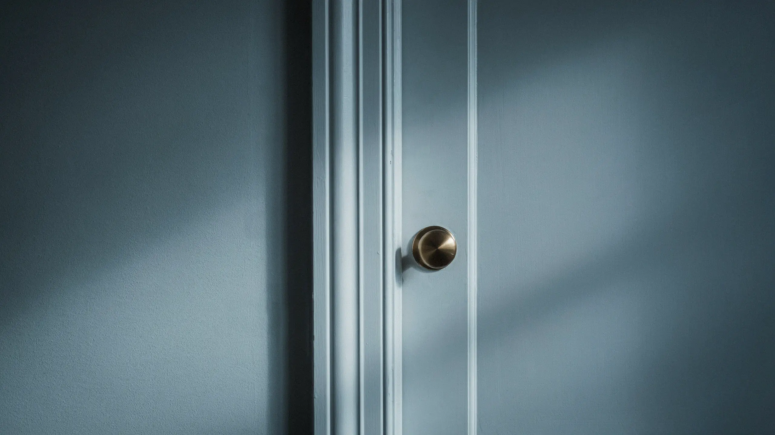 Close-up of a matte slate blue interior door featuring vertical decorative paneling and a round brushed brass doorknob, illuminated by soft directional window light that casts contrasting shadows across the wall and door surface.