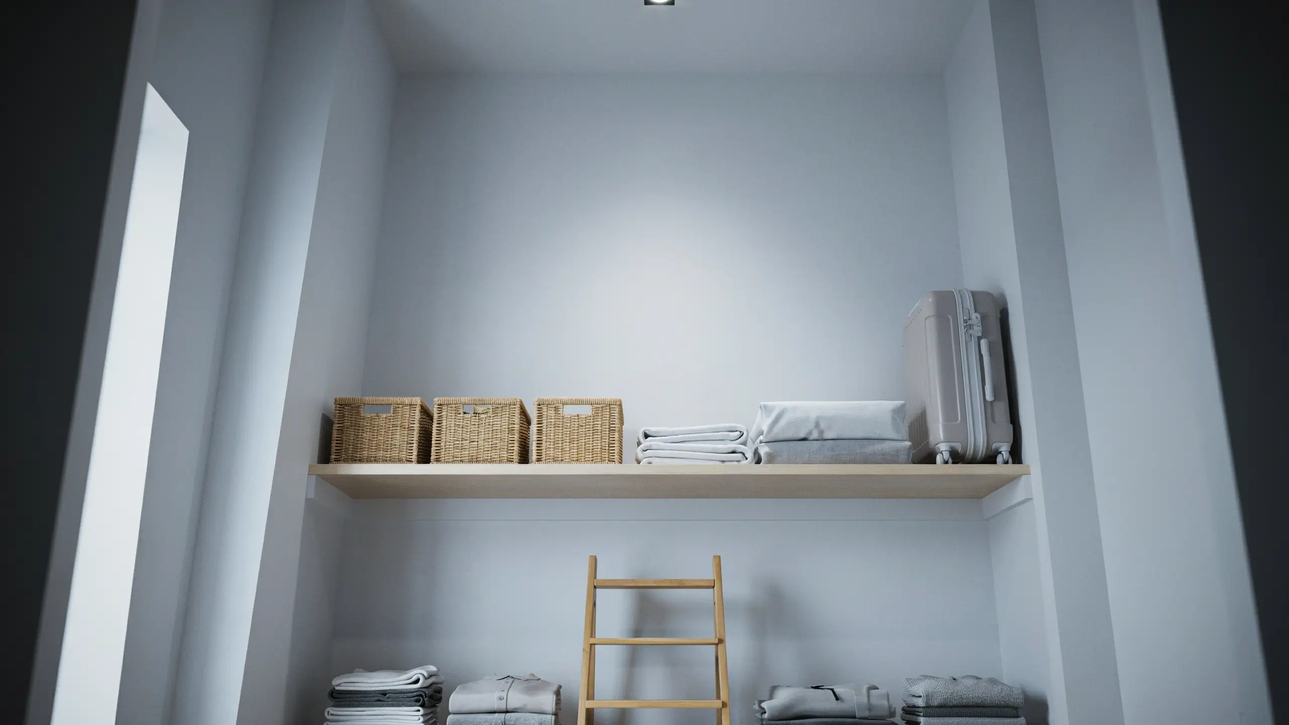 The main focal point is a light wood floating shelf installed high on the wall, holding three woven wicker storage baskets, a stack of folded white linens, and a light grey hardshell rolling suitcase. Below the shelf, a wooden ladder leans against the wall for easy access, and neat stacks of folded grey clothing rest on the floor, keeping the storage area organized and clutter-free.