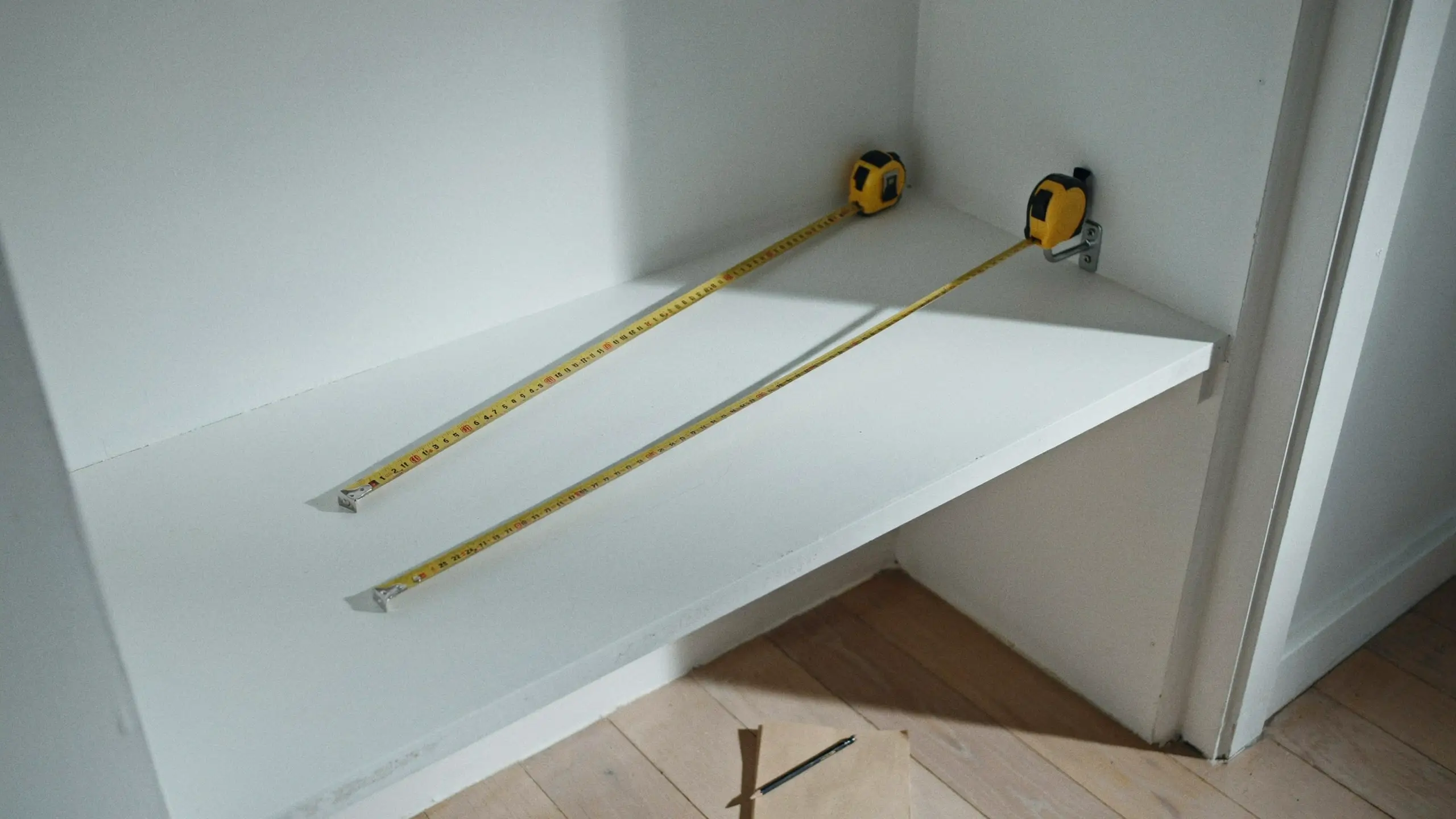 Close-up of a person using a yellow tape measure to measure the width of a wooden closet shelf before buying new storage bins.