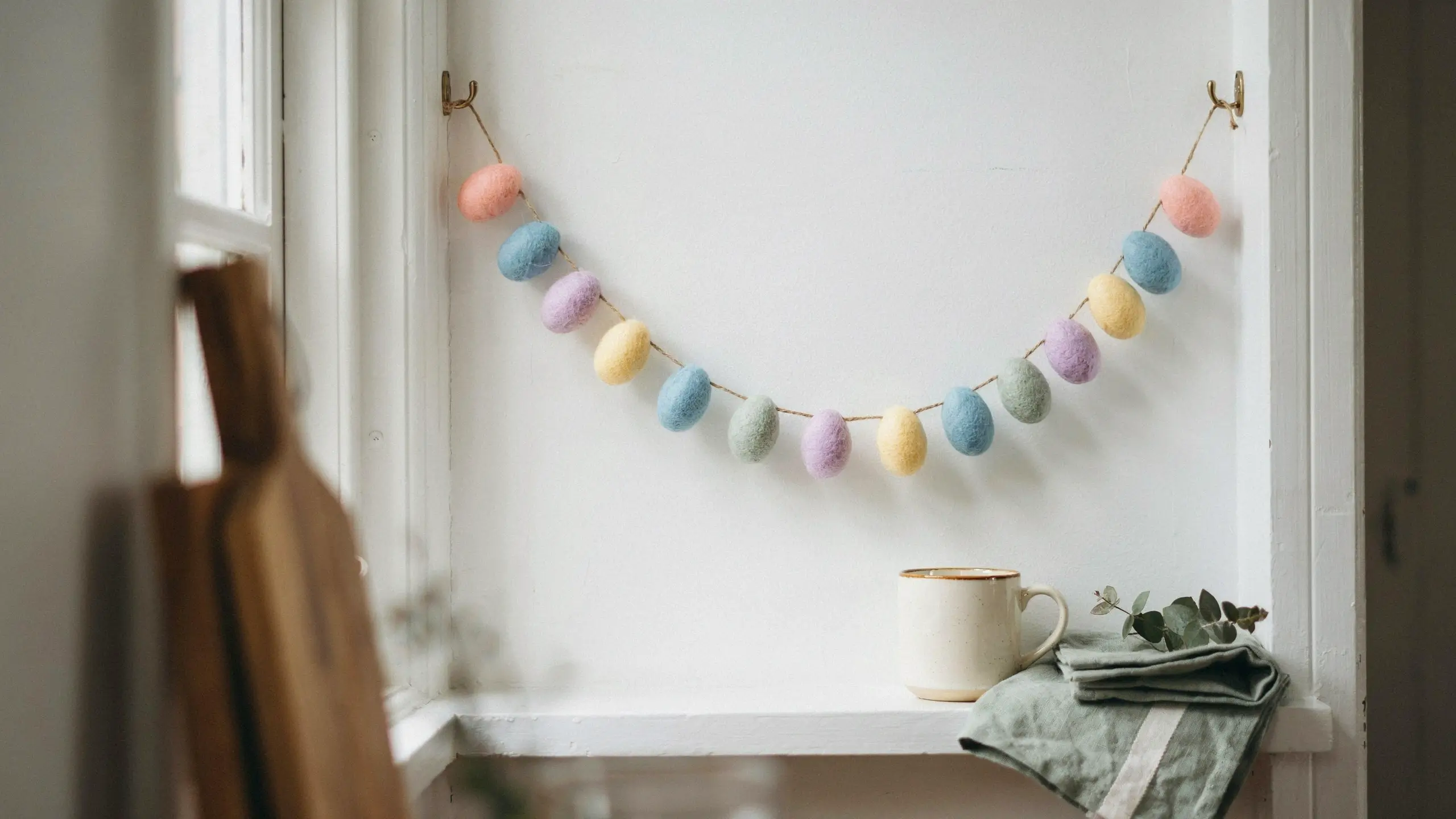 A pastel-colored mini felt egg garland hung from brass hooks across a white window niche, styled with a ceramic mug and folded sage linen cloth.
