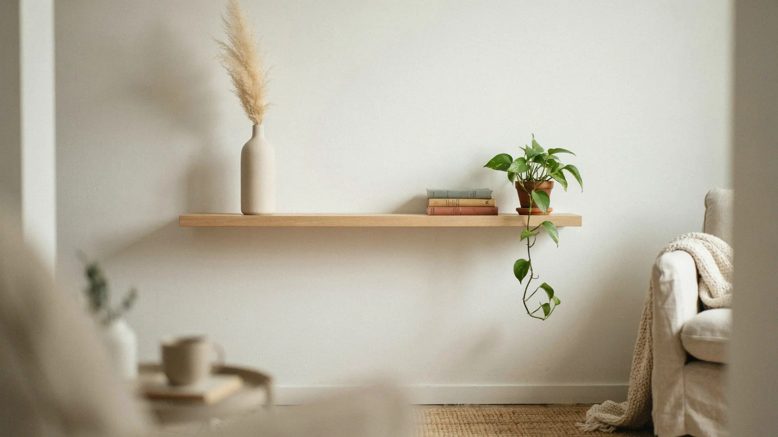 A warm neutral living room corner with a floating wooden shelf curated down to a few meaningful pieces: a tall cream vase holding fluffy pampas grass, a stack of vintage hardcover books, and a trailing green pothos plant beside a white armchair with a chunky knit throw blanket. 
