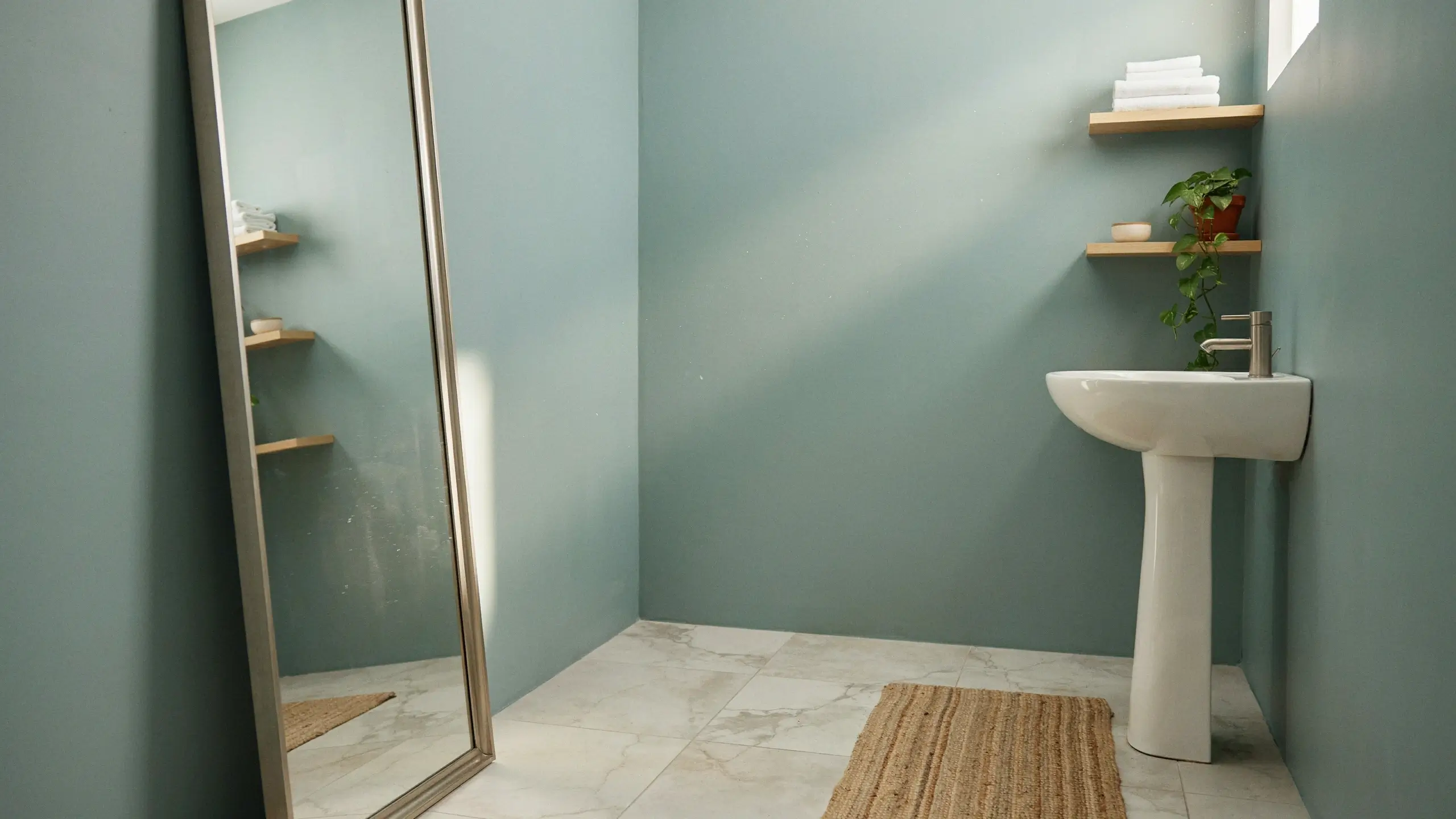 A tall, silver-framed standing mirror reflecting a stream of natural sunlight in a minimalist bathroom featuring a white pedestal sink, wooden corner shelves, a potted pothos plant, and clean marble floor tiles.