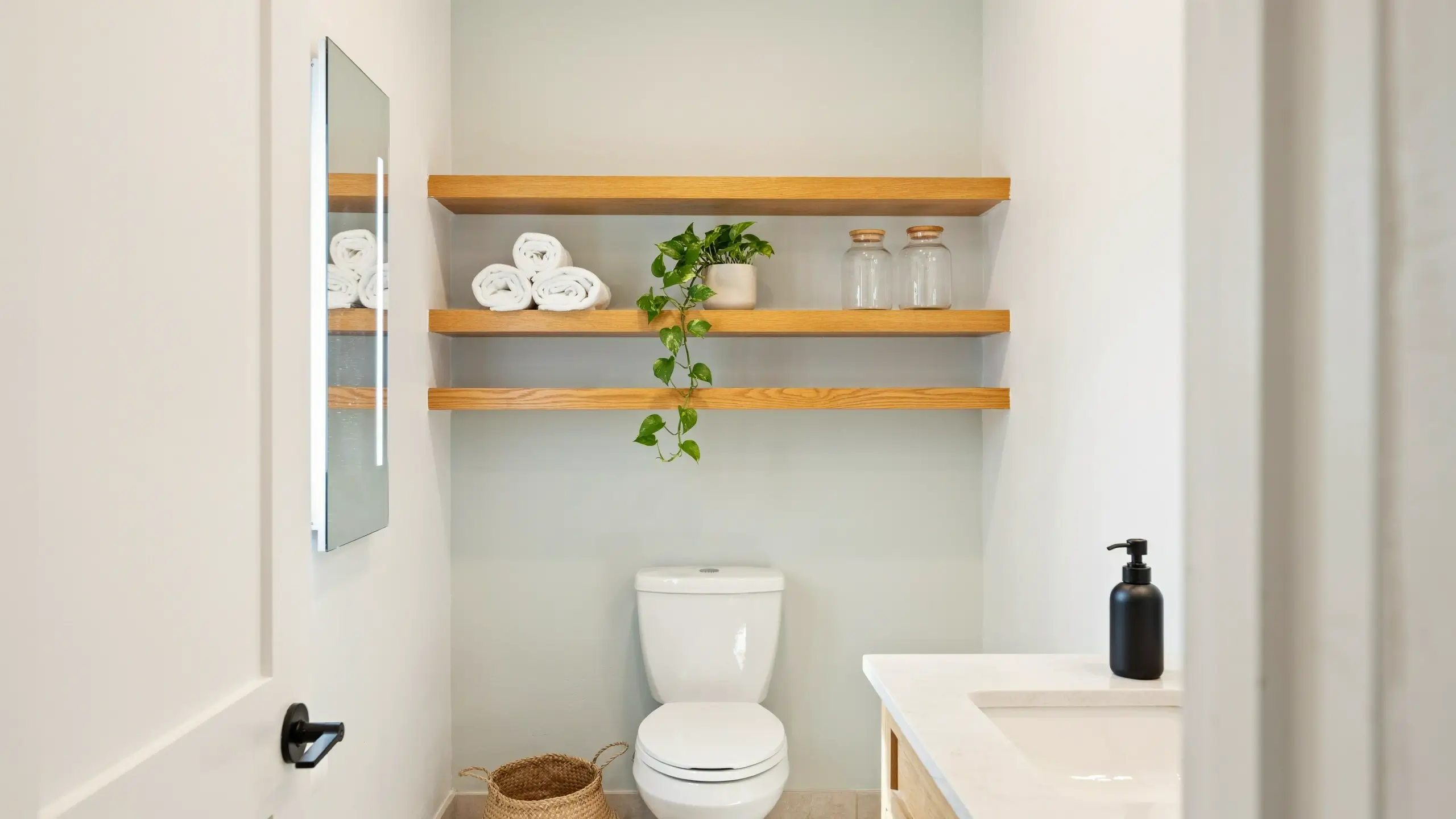 A clean, modern bathroom demonstrating wall-mounted storage: light wood floating shelves hold rolled white towels, glass jars, and a trailing green plant, keeping the sink counter completely clutter-free.