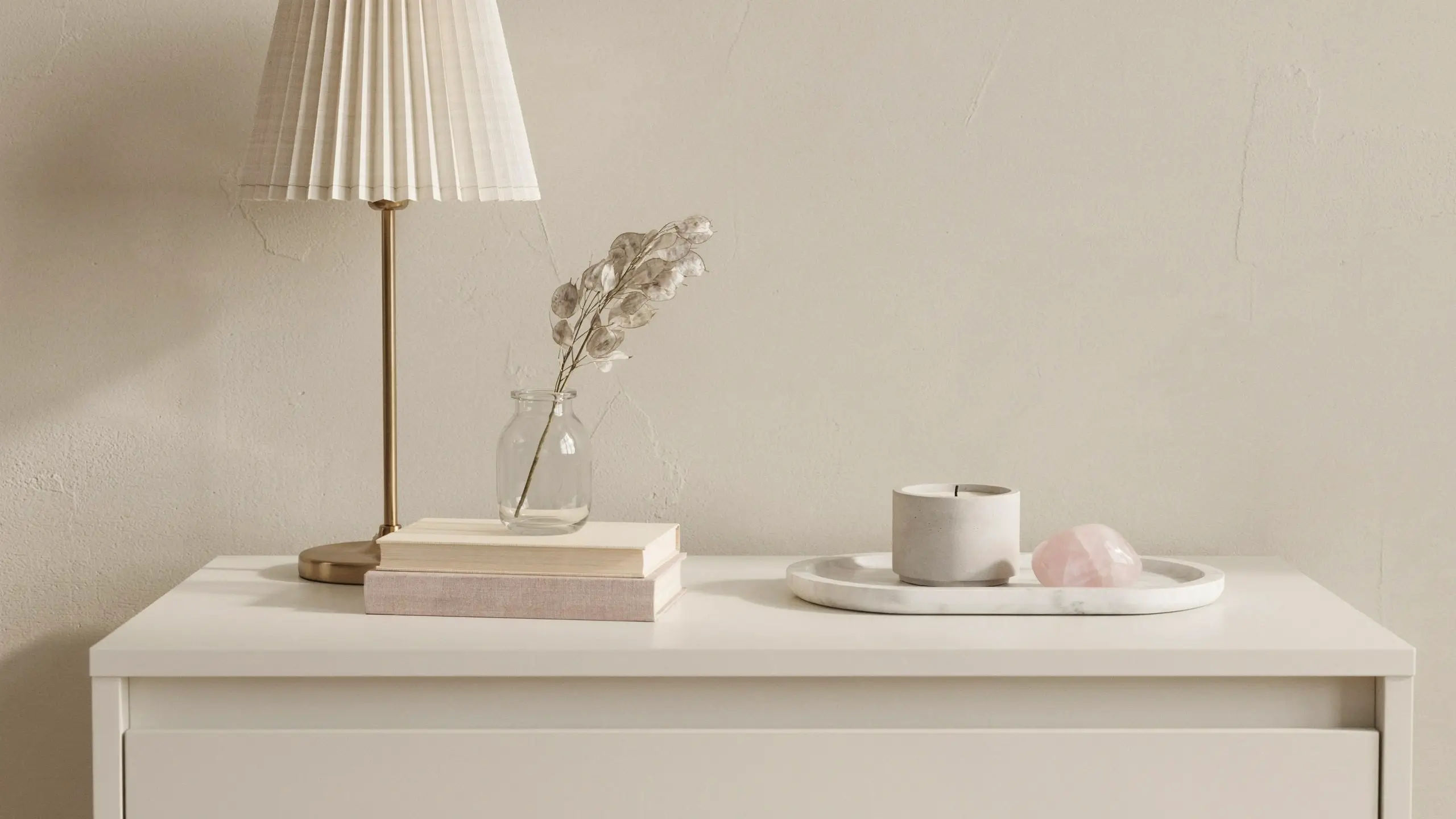 A minimalist white dresser styled with a cream pleated brass lamp, stacked books, dried cotton stems in a glass vase, a grey concrete candle, and a pink rose quartz crystal on a marble tray. 