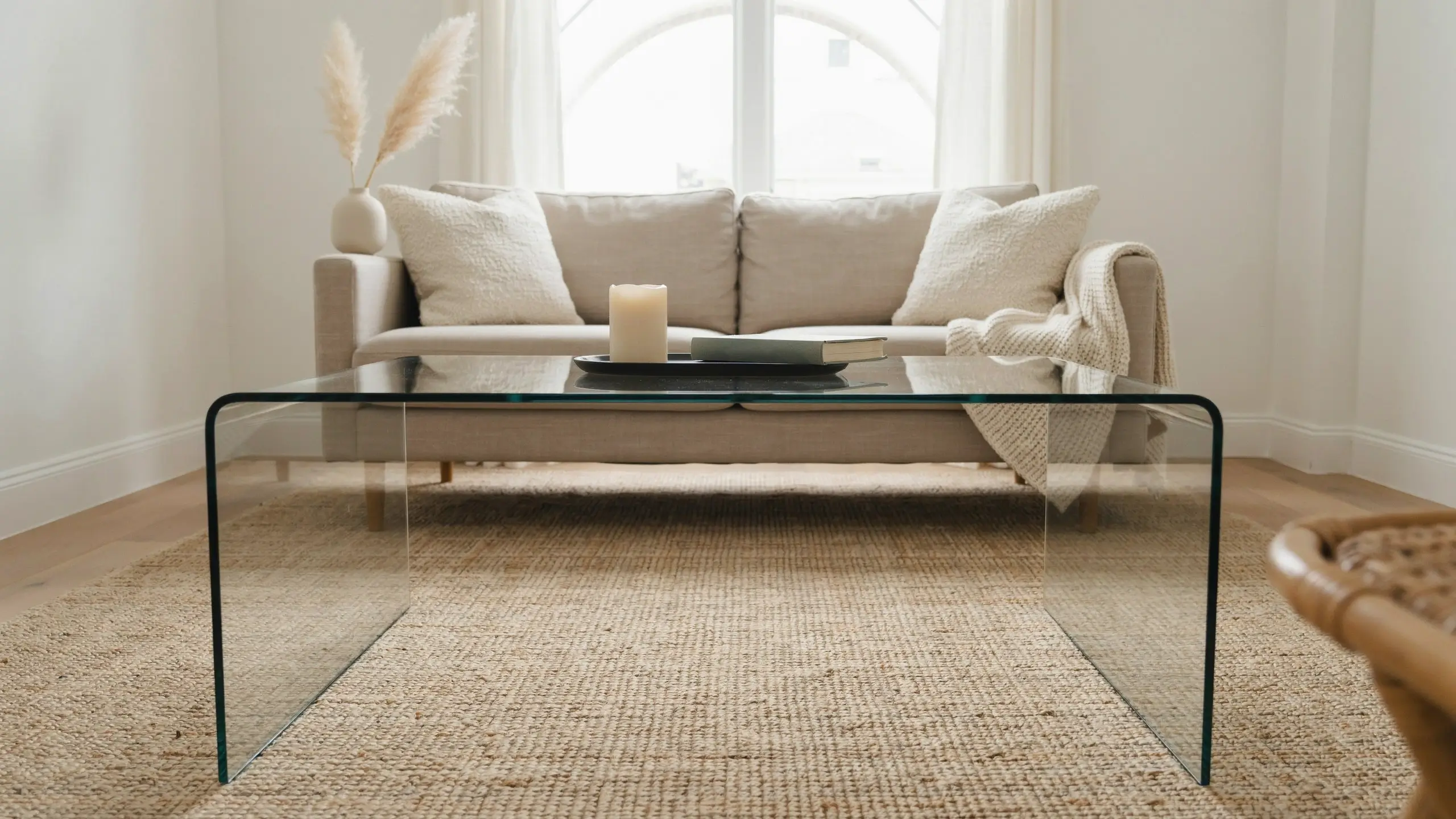 Minimalist bent glass coffee table in a neutral living room with a beige sofa, textured white pillows, pampas grass in a vase, and a cream throw blanket.