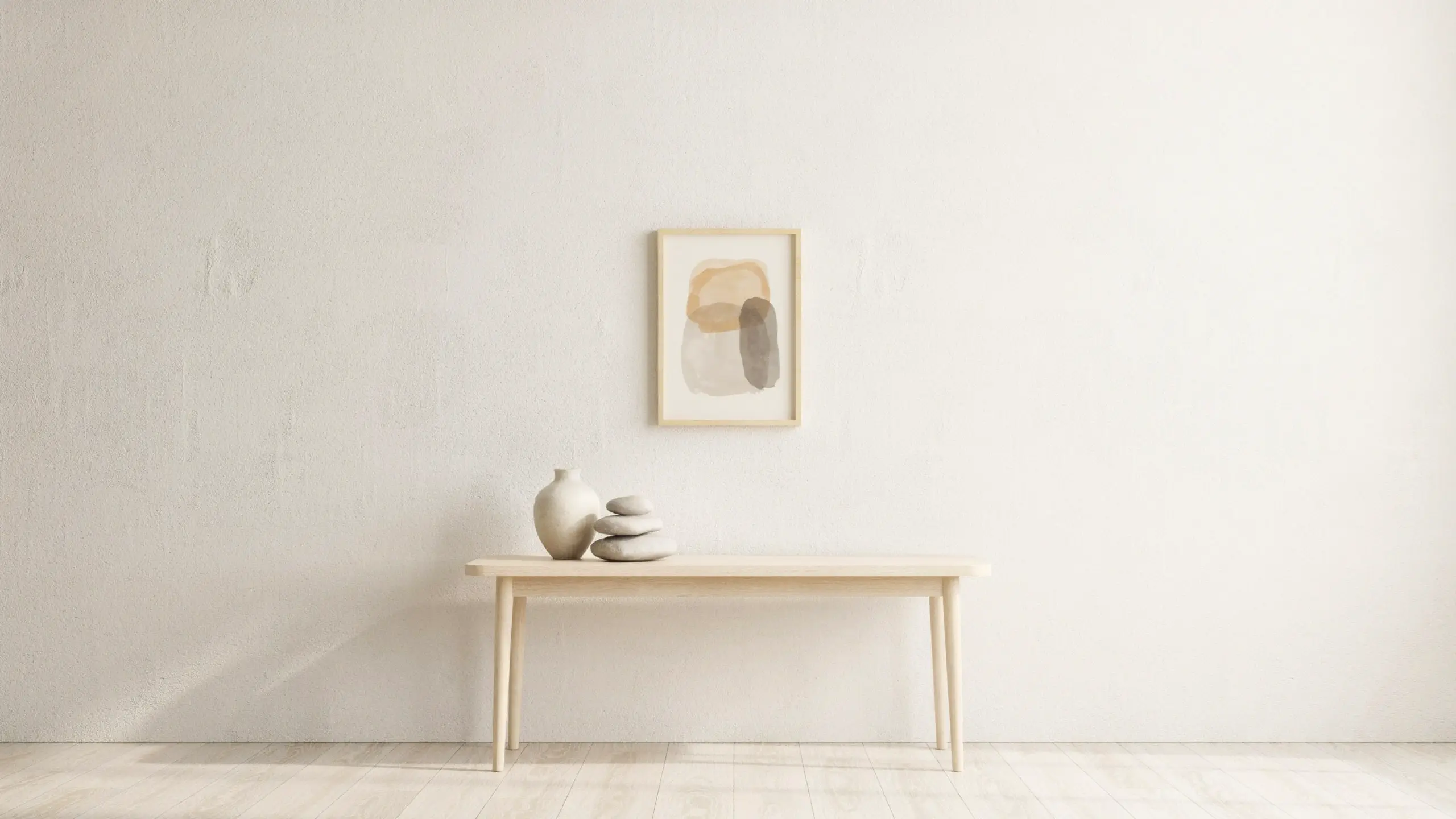 Minimalist light wood console table with a neutral vase, stacked zen stones, and framed abstract watercolor art on a textured off-white wall, showcasing negative space in home decor