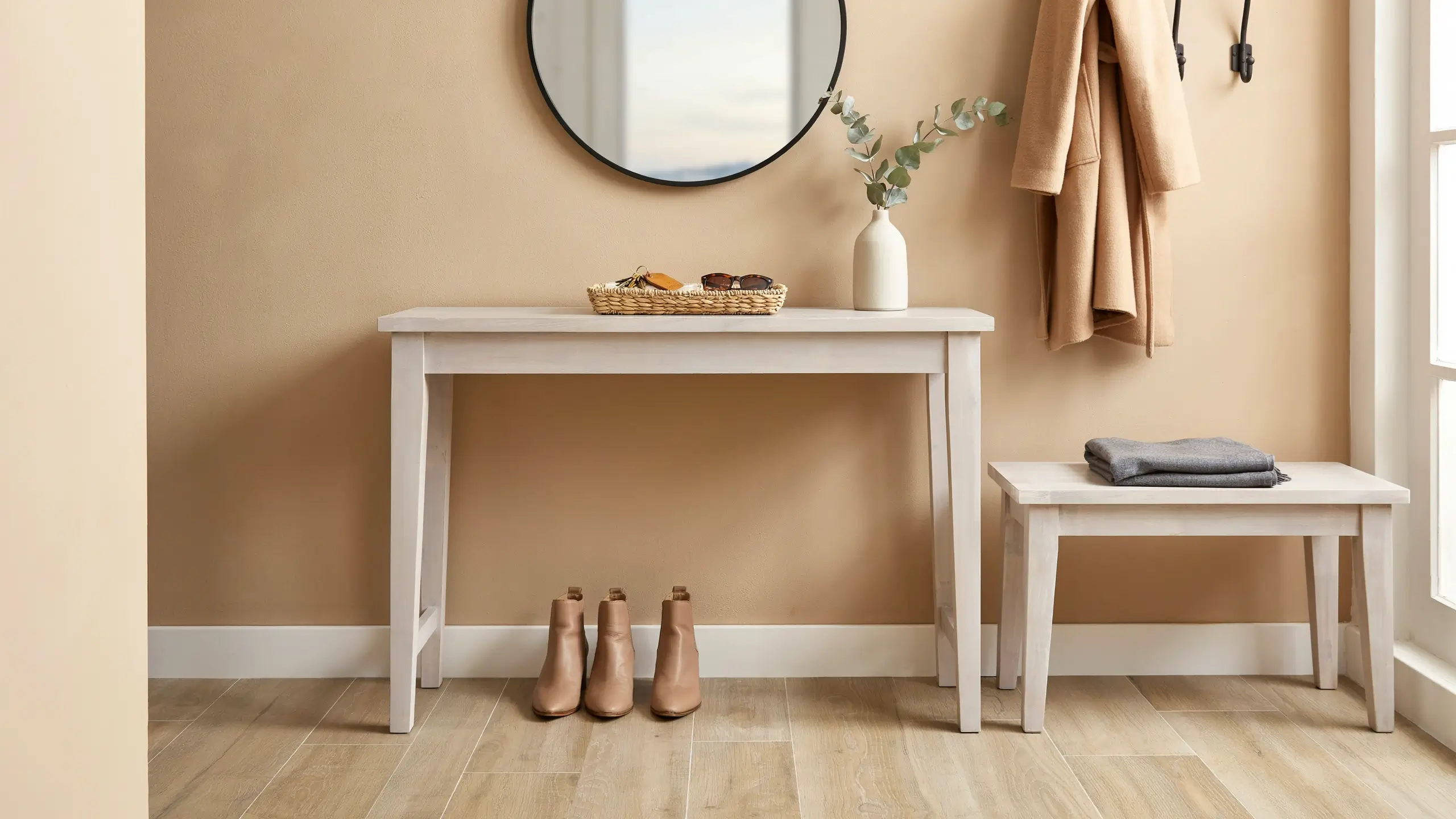 Minimalist white entryway with console table, round mirror, tan coat, eucalyptus vase, matching bench, and tan ankle boots on a wood floor.