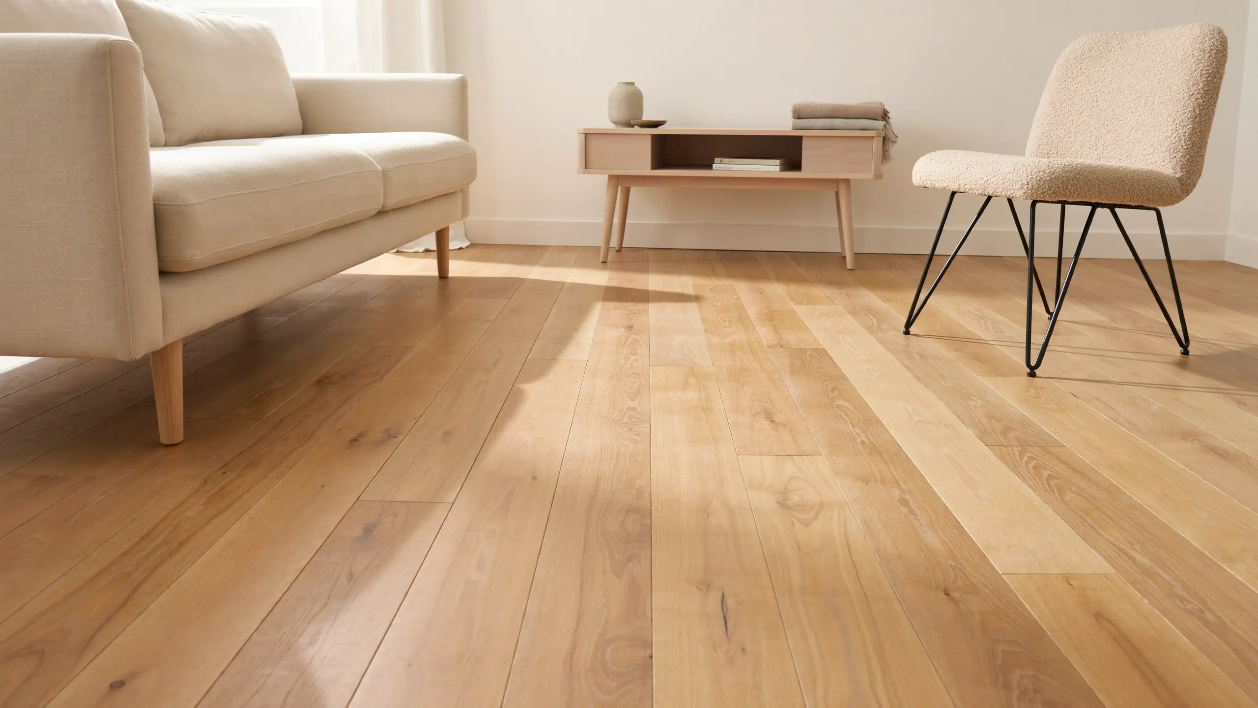 Minimalist living room with light oak wood flooring and furniture featuring visible space underneath, including a cream upholstered sofa with exposed light wood legs and a beige boucle accent chair with black metal hairpin legs.