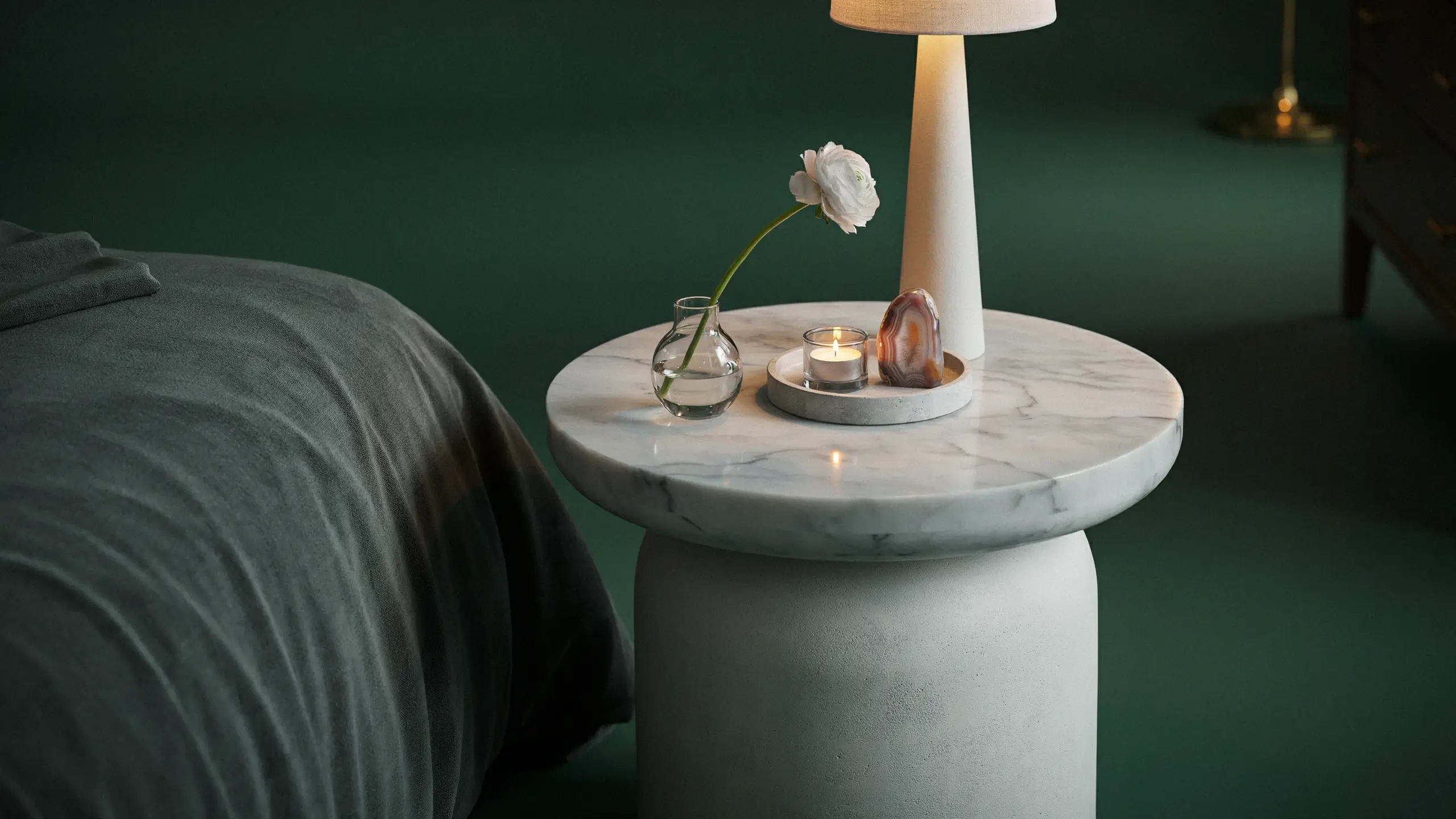 A minimalist round marble bedside table with a concrete pedestal base, holding a white conical lamp, a glass vase with a single white flower, and a lit candle on a small tray, set against a dark green wall in a modern bedroom interior. 
