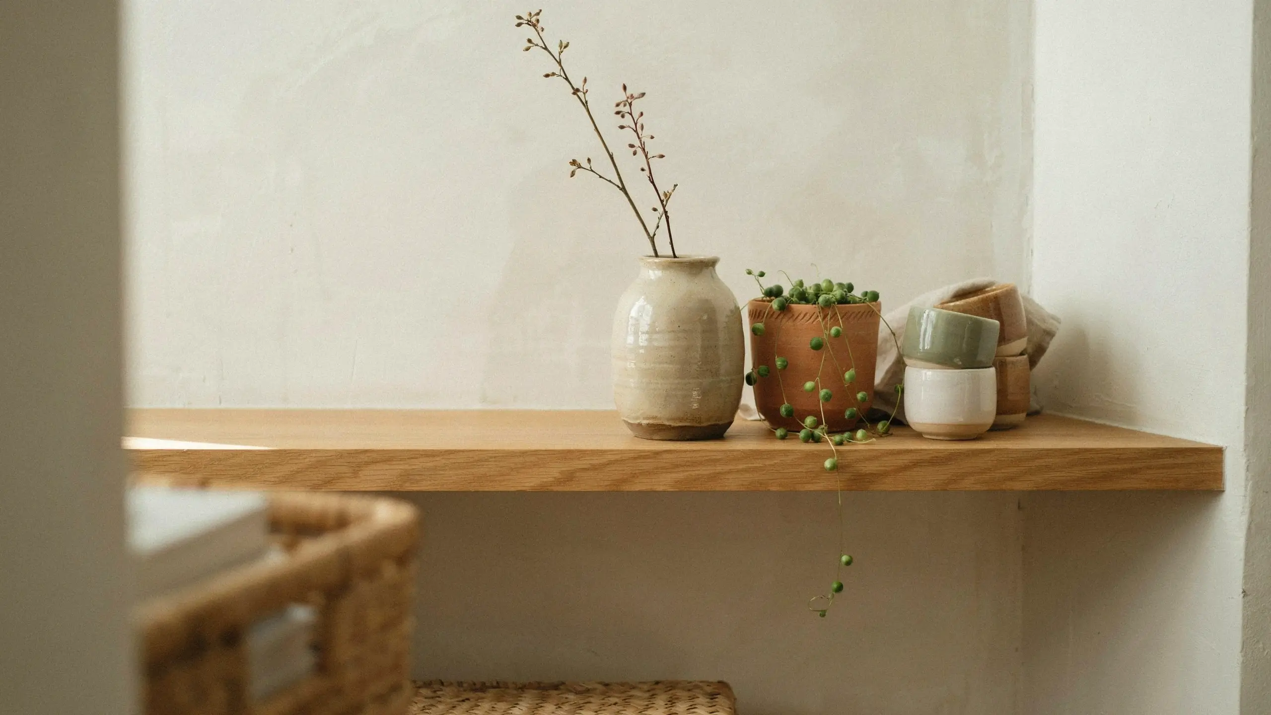 A minimalist wooden shelf styled with a beige glazed ceramic vase holding dried branches, a terracotta pot of trailing string of pearls, and stacked sage green and white handmade pottery cups with a folded linen cloth