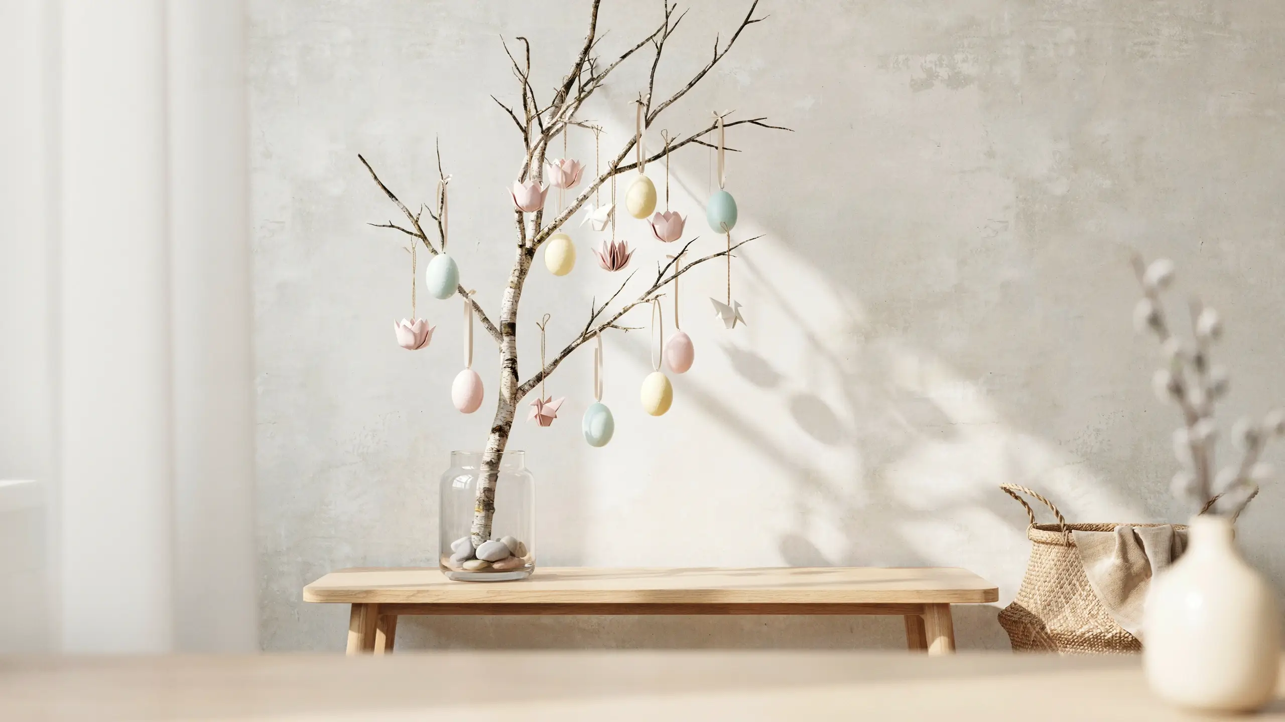 Minimalist spring tree branch display featuring pastel Easter eggs and flower ornaments in a glass jar with river stones on a light wooden bench.