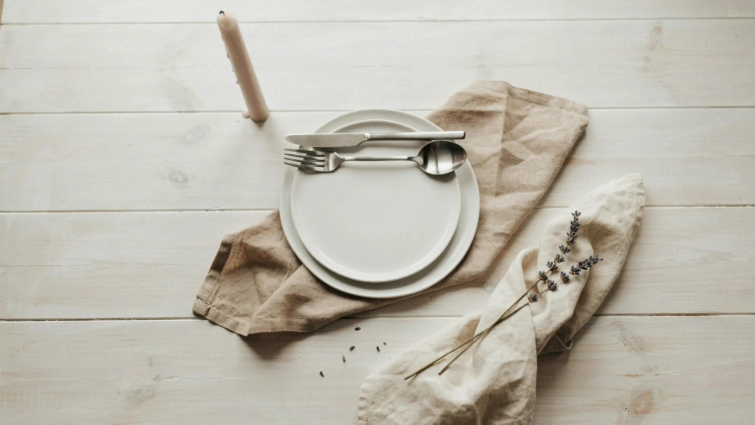White minimalist place setting featuring tea towels used as table linens under stacked plates, with silverware and a dried lavender sprig