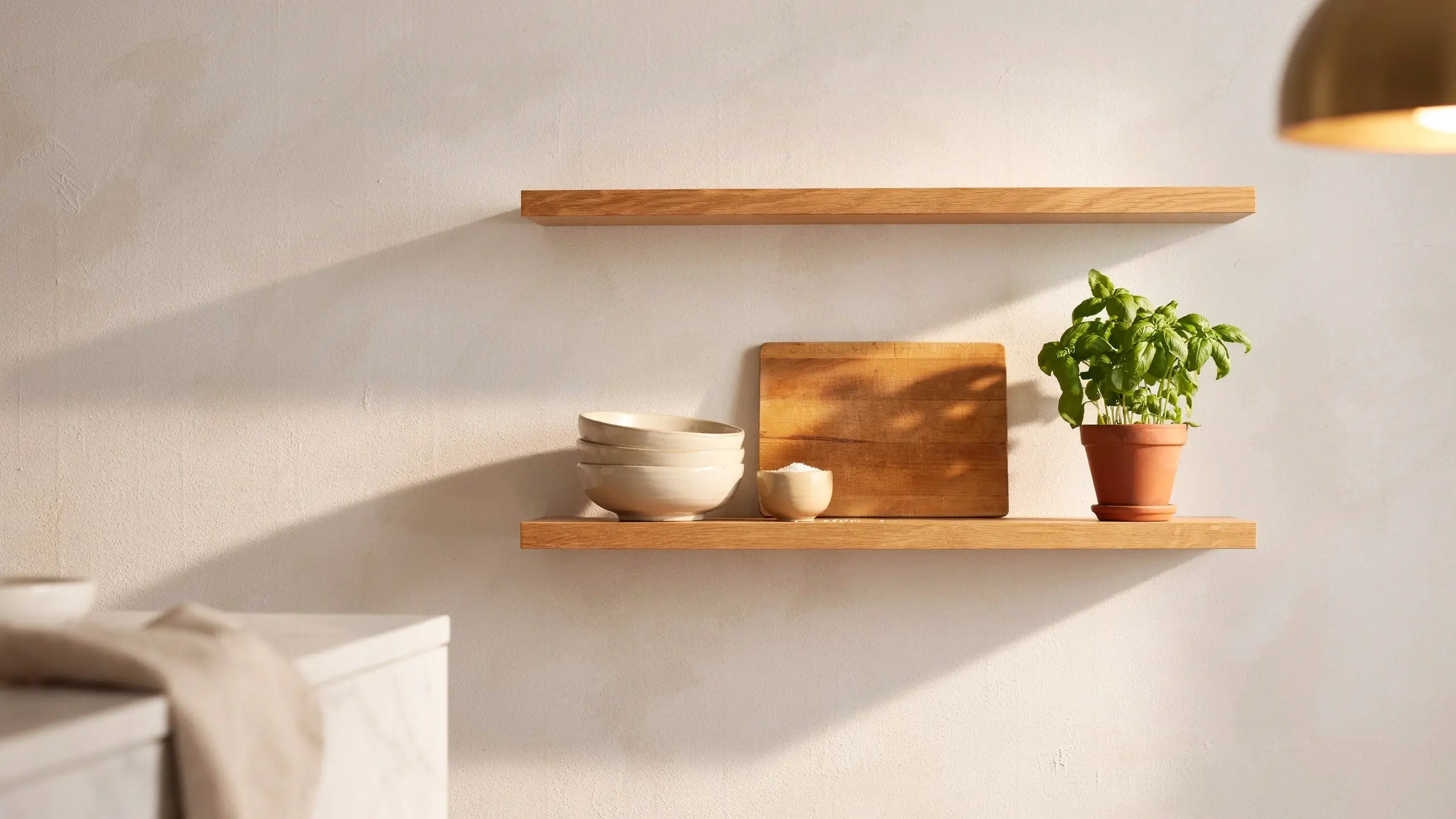 Two light wooden floating shelves mounted on a textured white wall, featuring stacked cream ceramic bowls, a wooden cutting board, and a potted basil plant illuminated by warm sunlight.