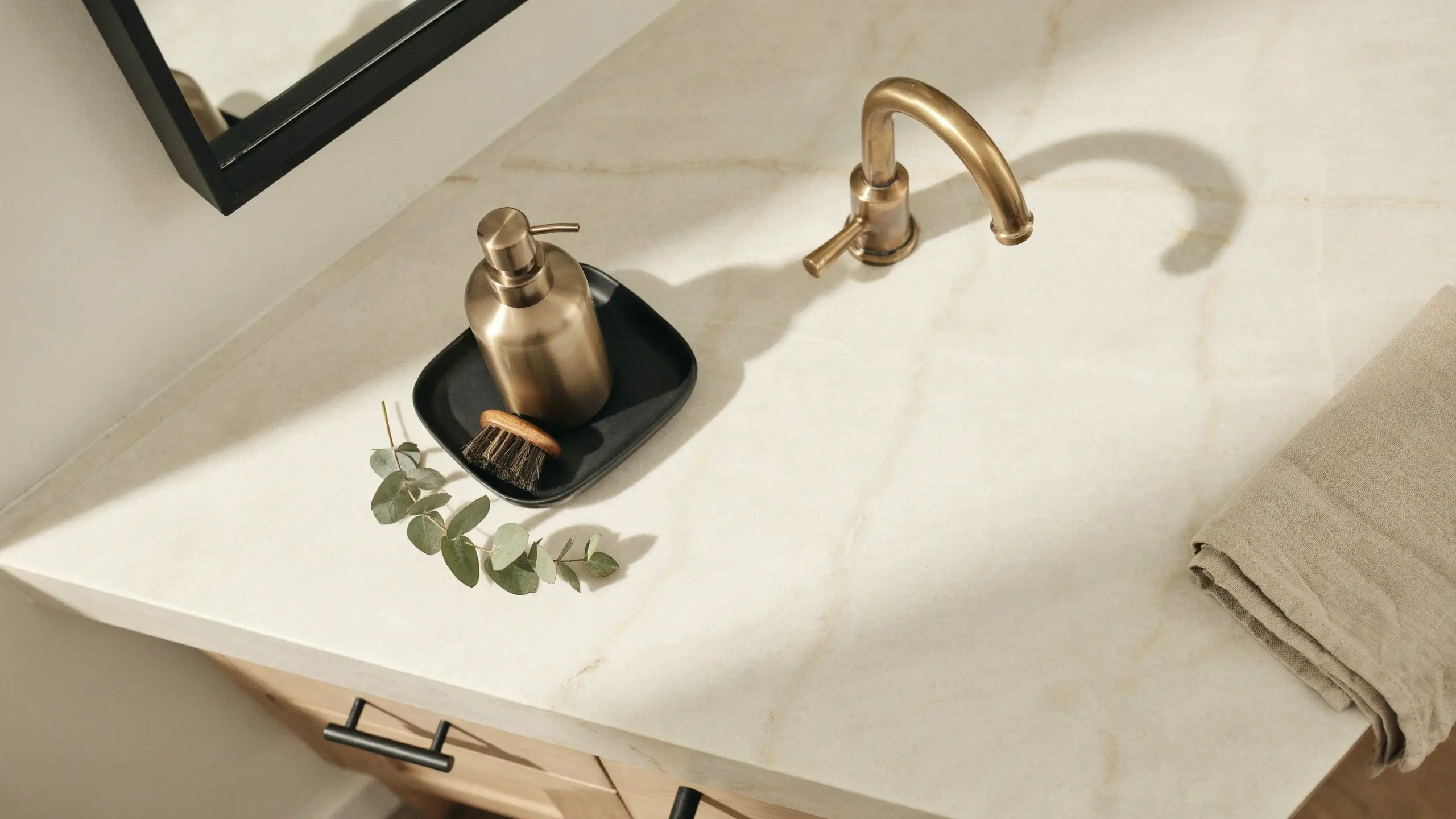 A minimalist bathroom vanity top with an antique brass faucet and matching soap dispenser placed on a matte black tray. A wooden hand scrub brush, fresh green eucalyptus sprig, beige linen towel, and black-framed mirror sit on the white marble counter, demonstrating the interior design trend of mixing two metals instead of five.