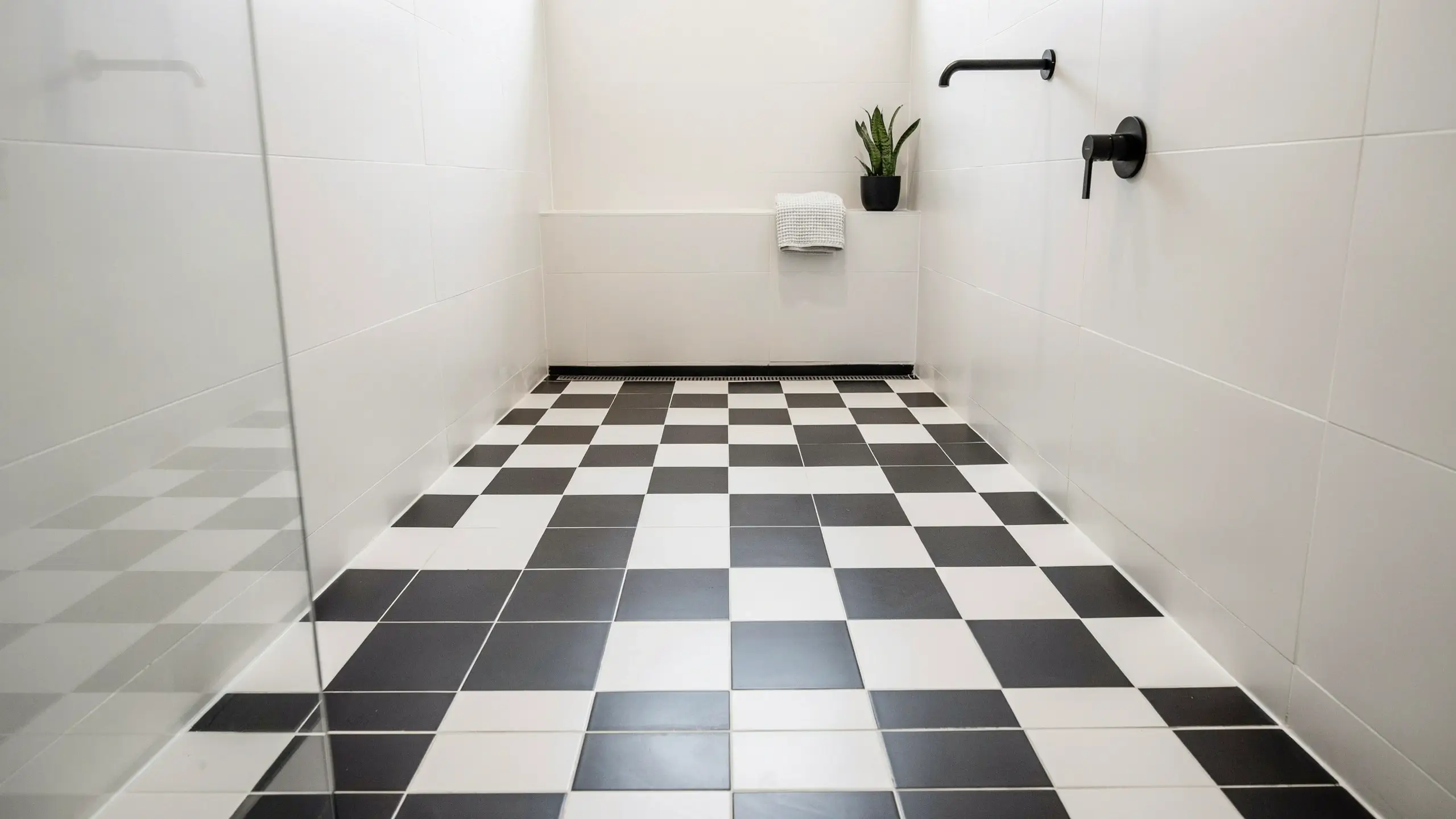 Black and white checkerboard floor tiles in a modern walk-in shower with matte black fixtures, white wall tiles, and a linear drain.