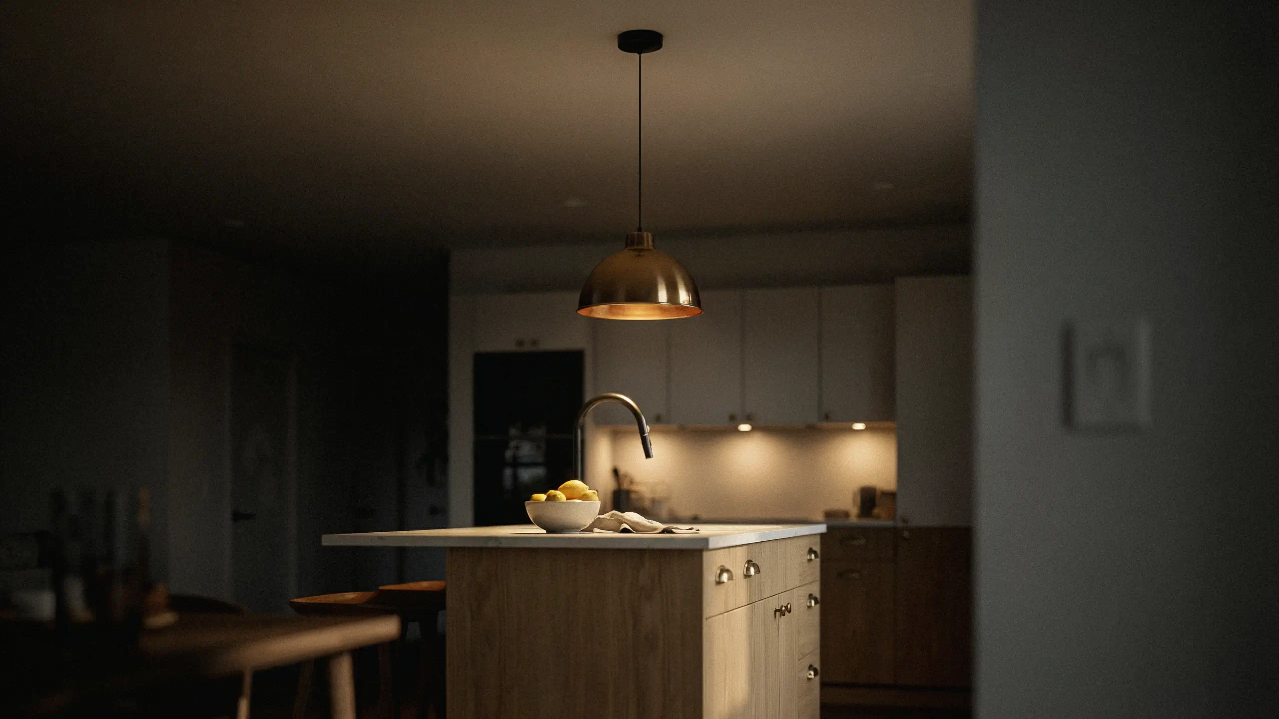 A cozy kitchen island illuminated by a warm brass pendant light, featuring a white countertop, wooden paneled base, high-arc faucet, and a bowl of fresh lemons for an inviting, ambient atmosphere.