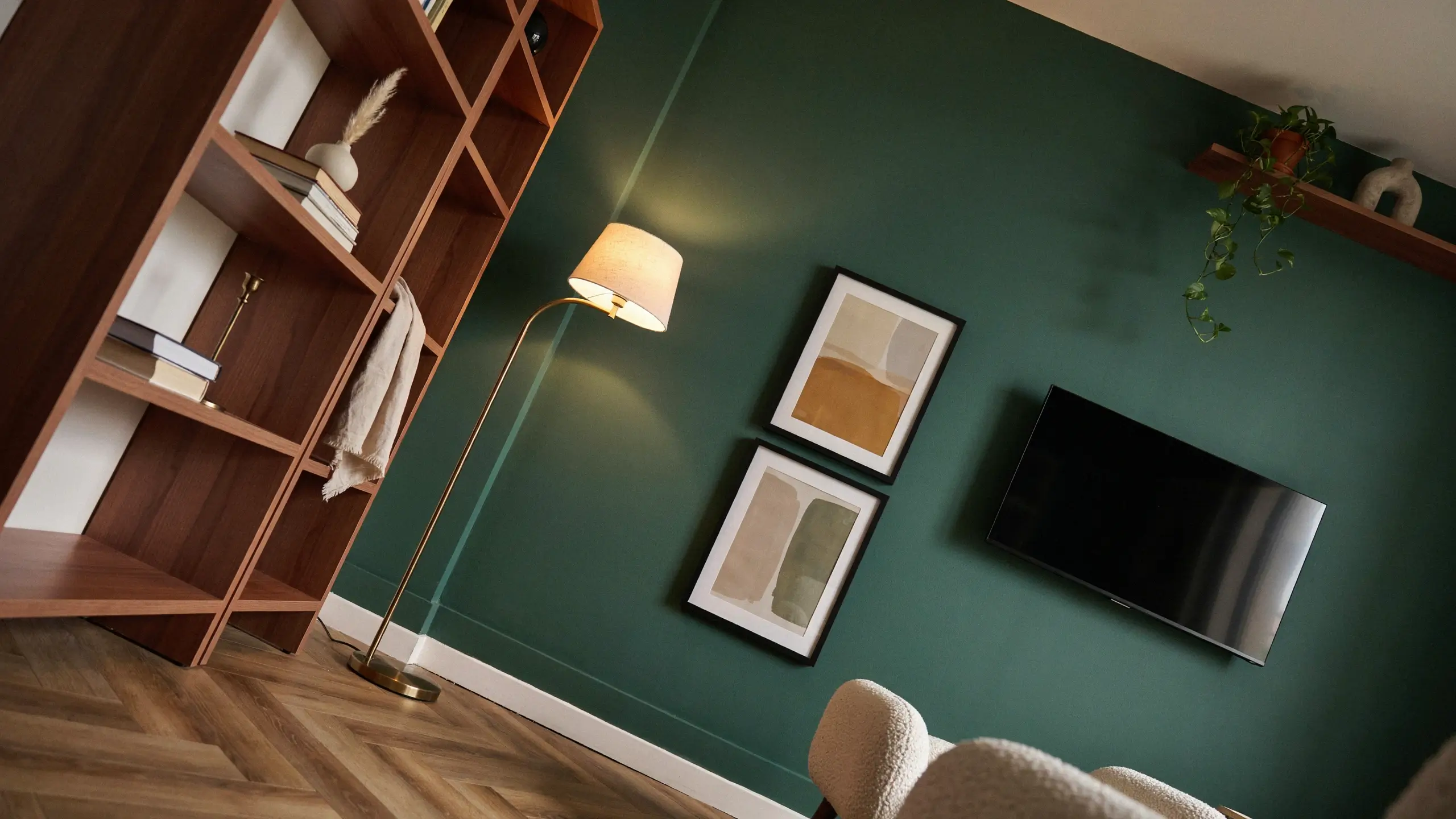 Modern living room interior featuring a dark green accent wall, wooden open shelving unit filled with books and decorative items, a brass floor lamp casting a warm glow over two framed abstract art prints, a wall-mounted flat-screen television, and a white boucle armchair in the foreground.