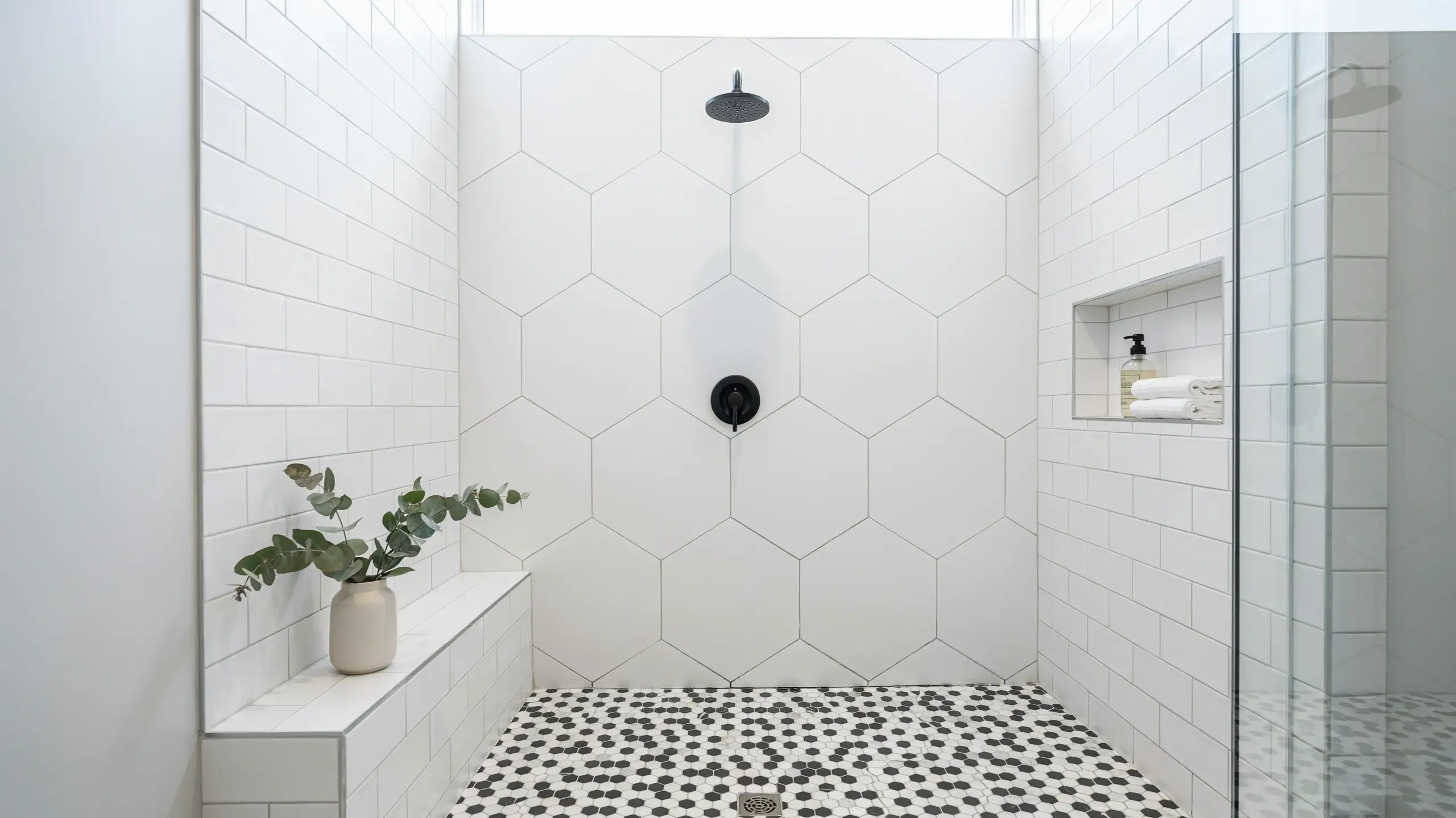 Modern bathroom shower interior with a large white hexagon tile accent wall, matte black rainfall showerhead and control valve, white subway tile sides, a recessed niche holding soap and folded towels, a white built-in bench with a eucalyptus plant, and a black-and-white penny mosaic tile floor with a square drain.
