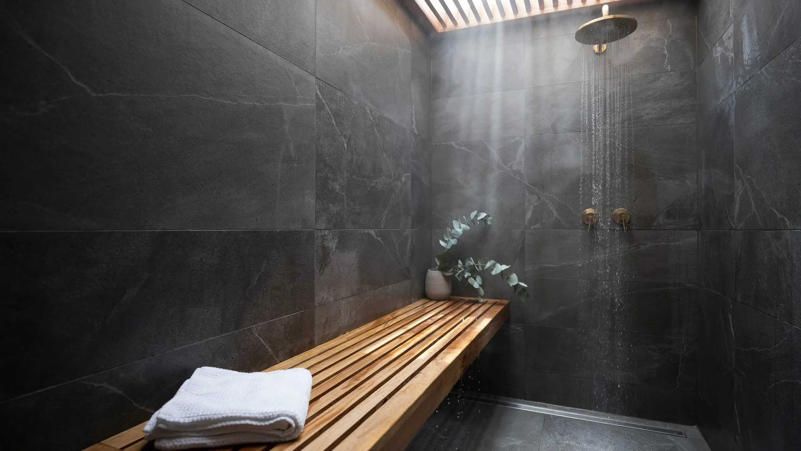 A luxury walk-in shower with brass rainfall fixtures and a natural wood slatted bench holding a folded white towel and potted eucalyptus, warming up the cool stone tile walls.
