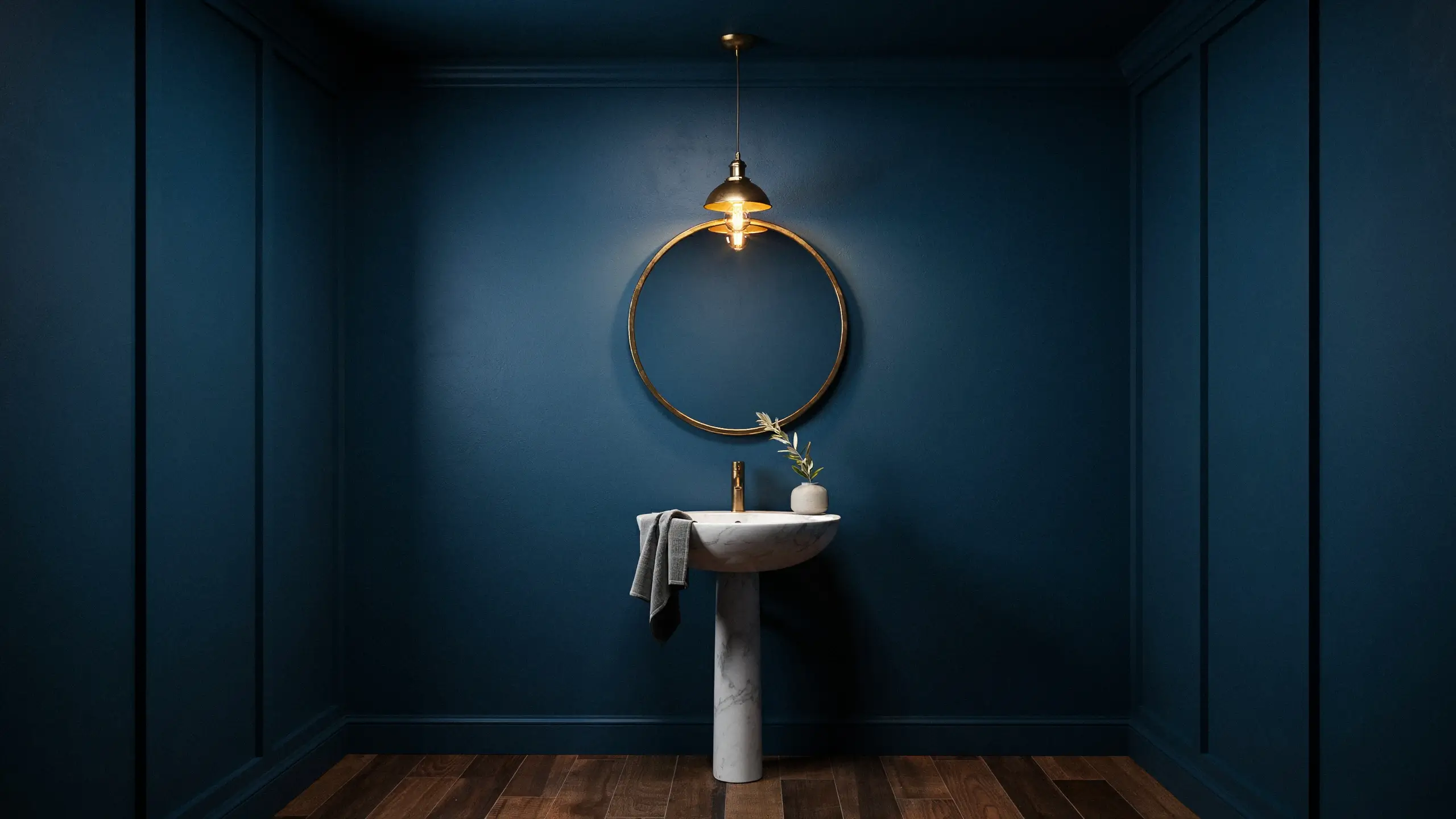 Dark blue moody bathroom with a white marble pedestal sink, gold-framed round mirror, and warm pendant lighting.
