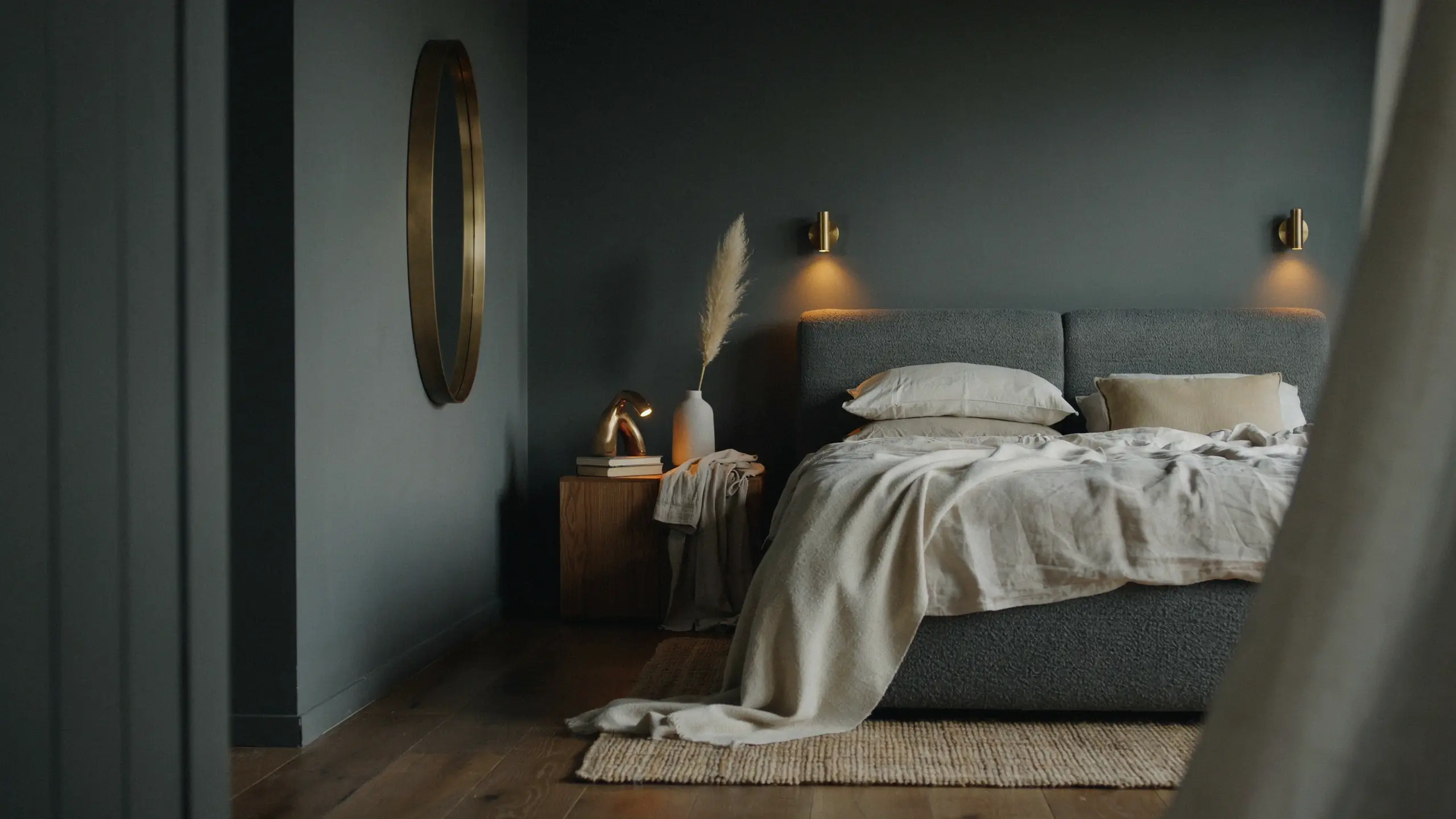 Moody bedroom interior with dark charcoal walls, a brass oval mirror, warm wall sconces, and a grey upholstered bed dressed in cream linen bedding and a textured throw blanket, featuring a wooden nightstand with brass lamp and pampas grass.