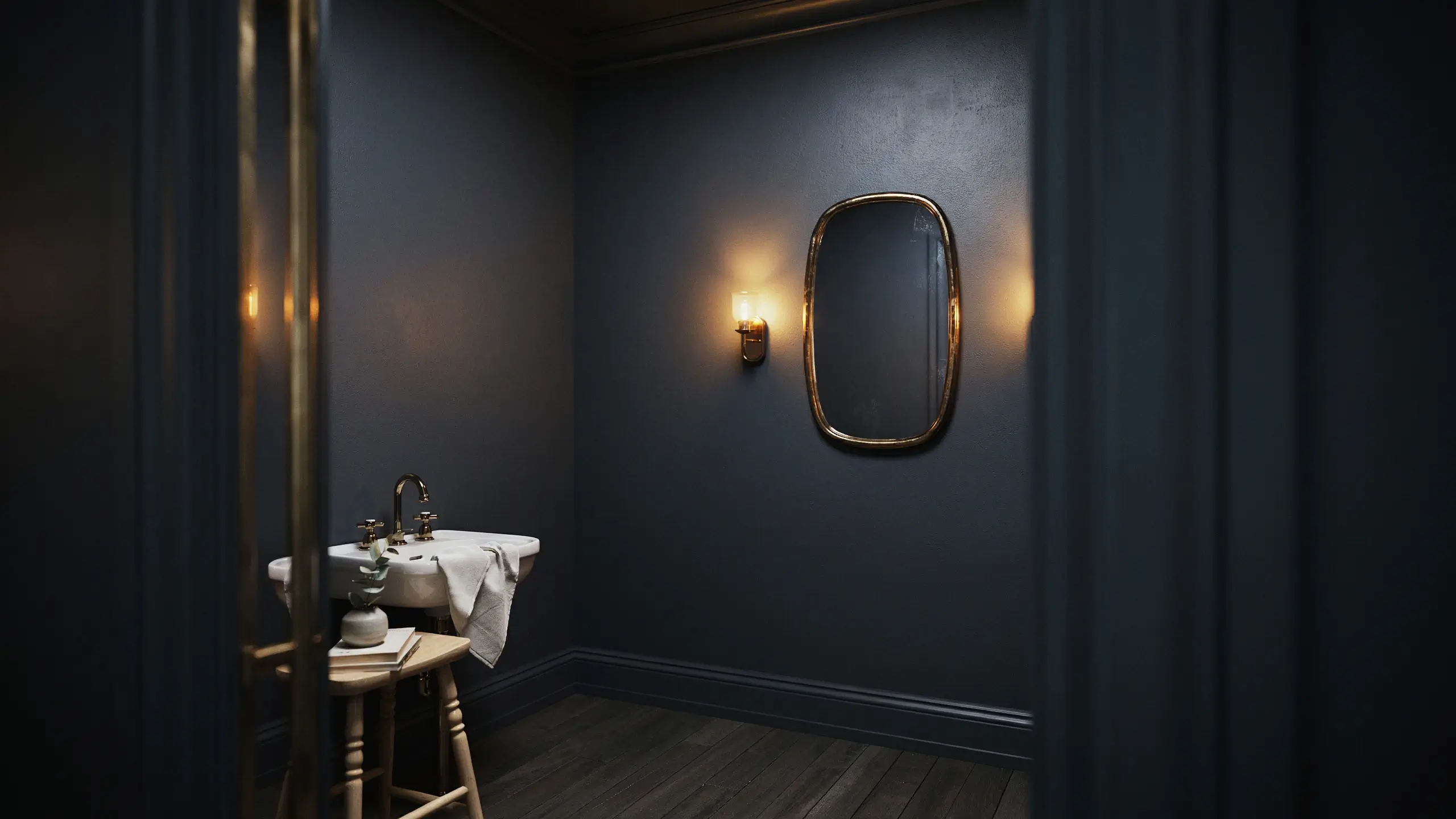 Dark moody bathroom with slate blue walls, a rounded brass mirror, warm wall sconce, and white porcelain sink on a wooden stool.
