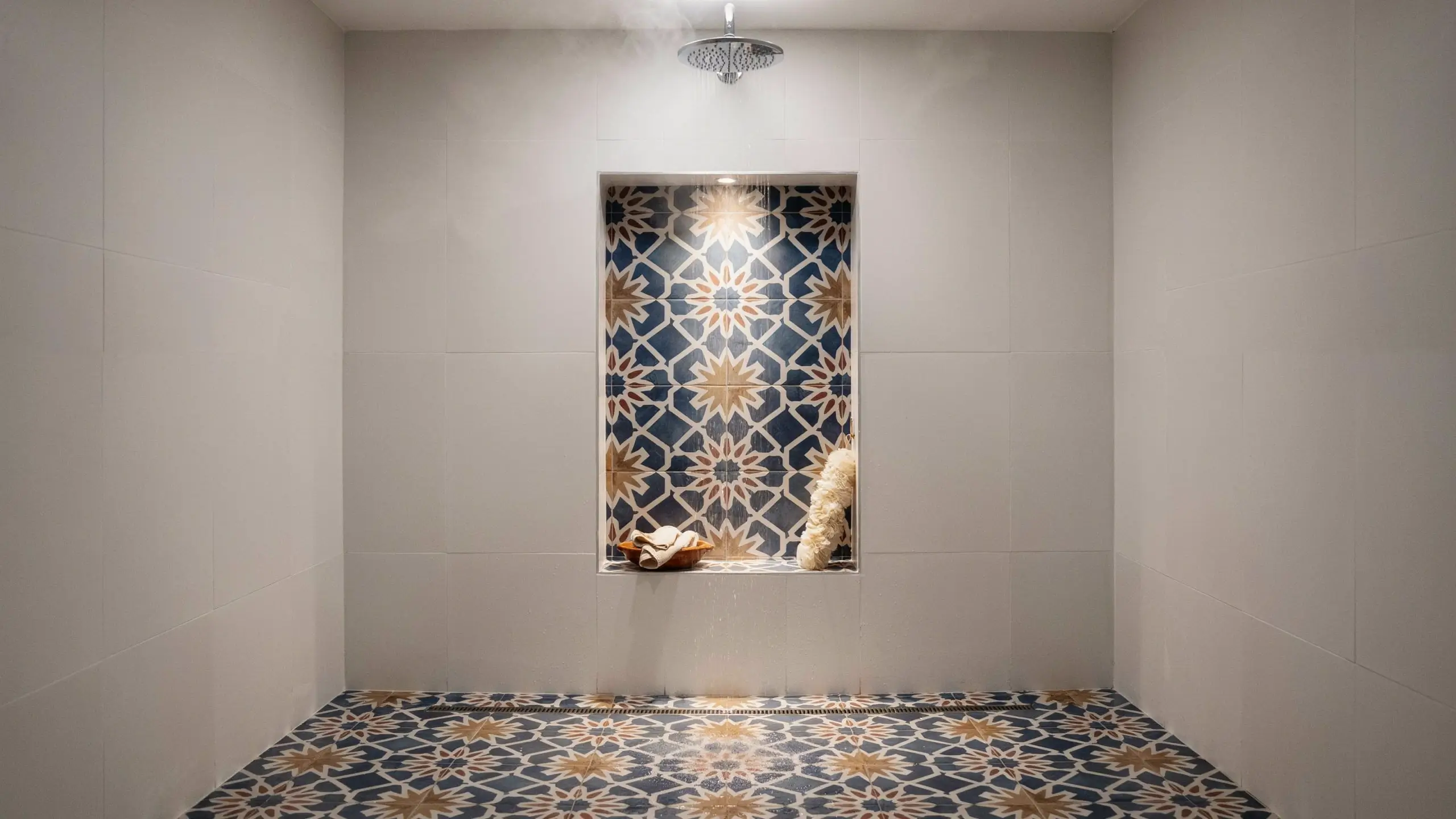Modern walk-in shower featuring a rainfall showerhead and a recessed wall niche with a blue, beige, and white geometric Moroccan-inspired pattern tile, holding a natural loofah and a wooden bath sponge.