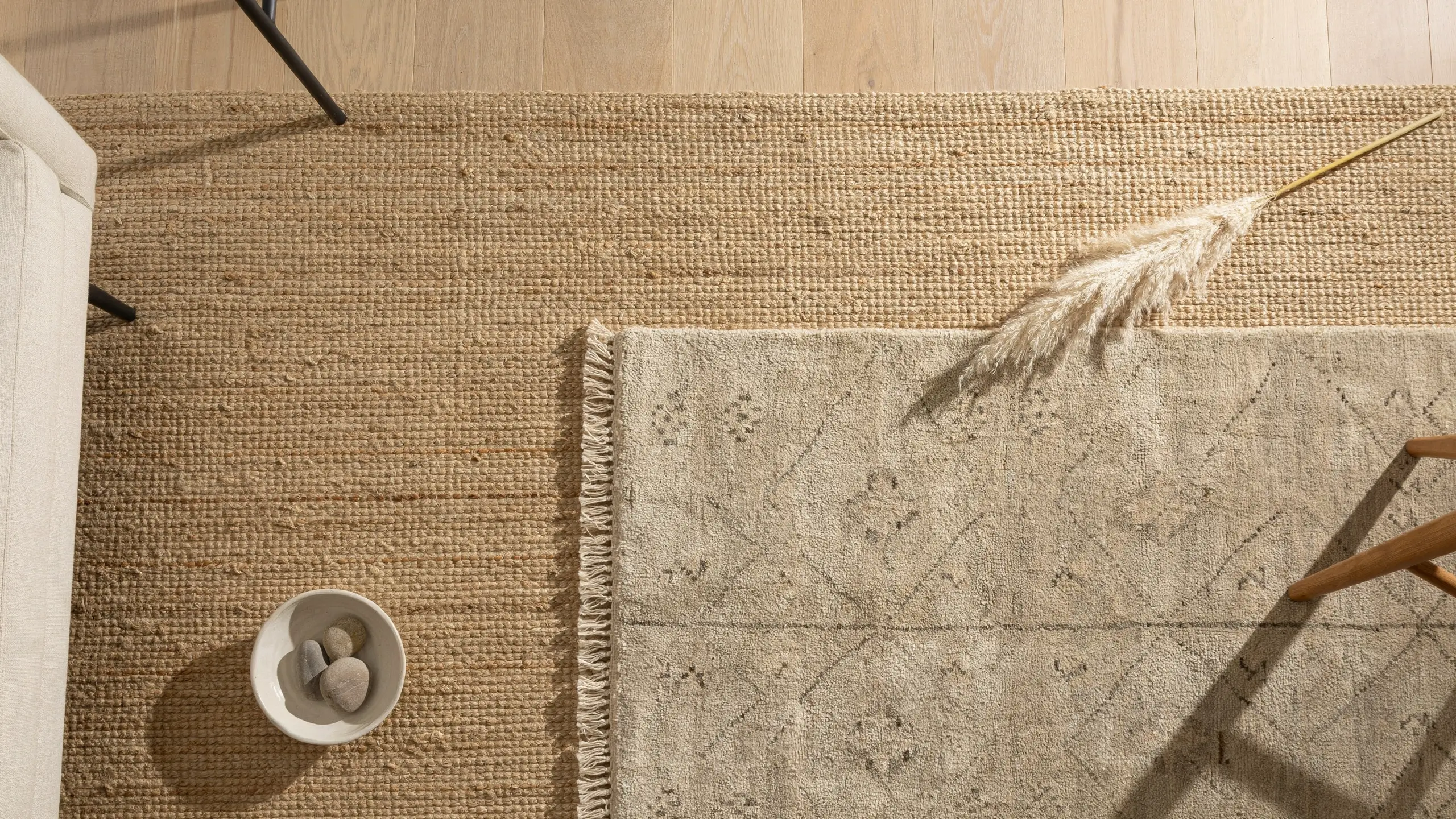 Overhead flat lay of a woven natural fiber jute rug layered with a patterned fringe textile, a minimalist white bowl of smoothing stones, and dried pampas grass, illustrating the "ground the room" interior styling principle.
