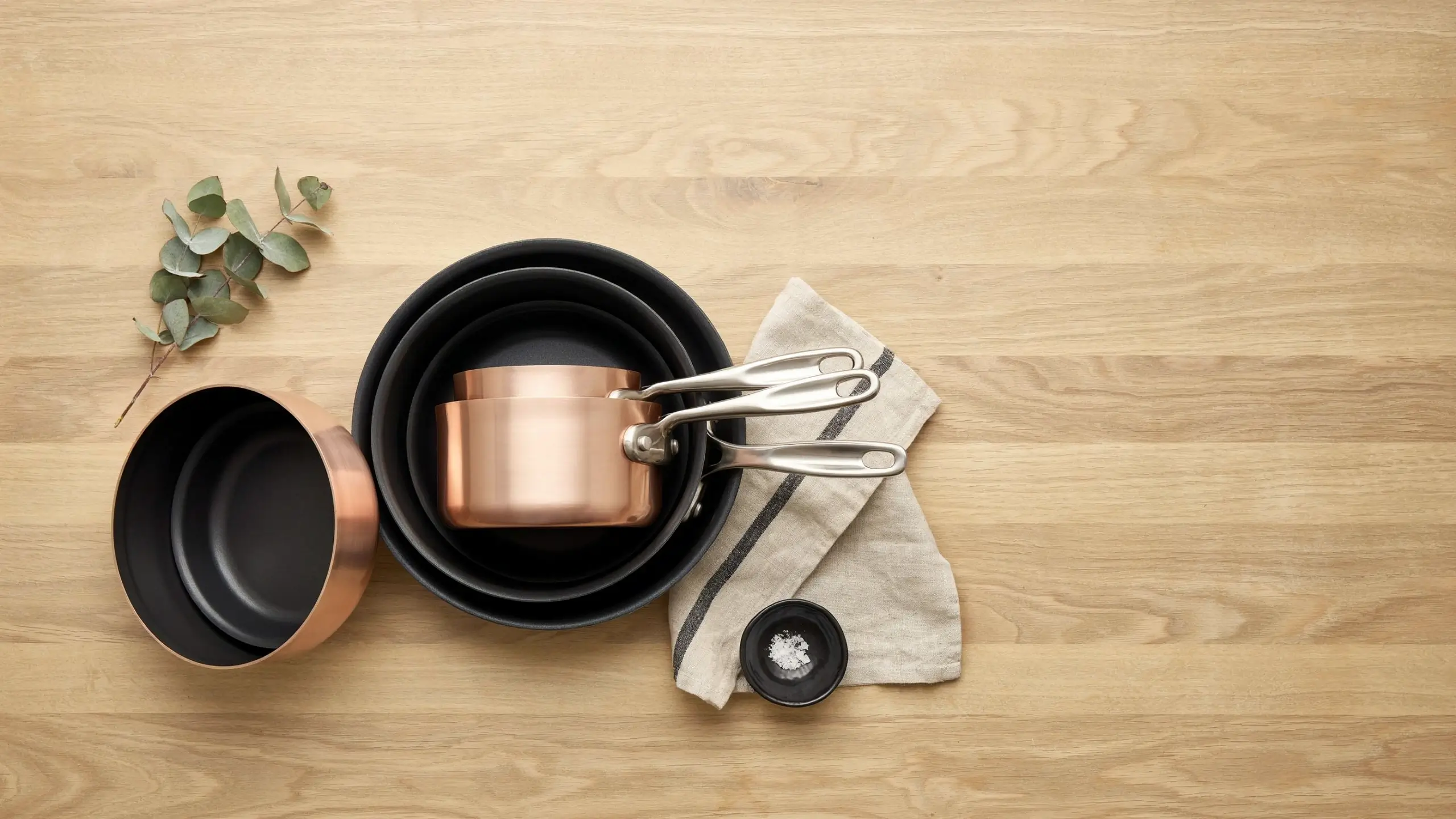 A stack of nested black frying pans and a polished copper saucepan with stainless steel handles, arranged on a light wooden tabletop with a sprig of eucalyptus, a striped linen cloth, and a small bowl of coarse sea salt for a minimalist kitchen aesthetic.