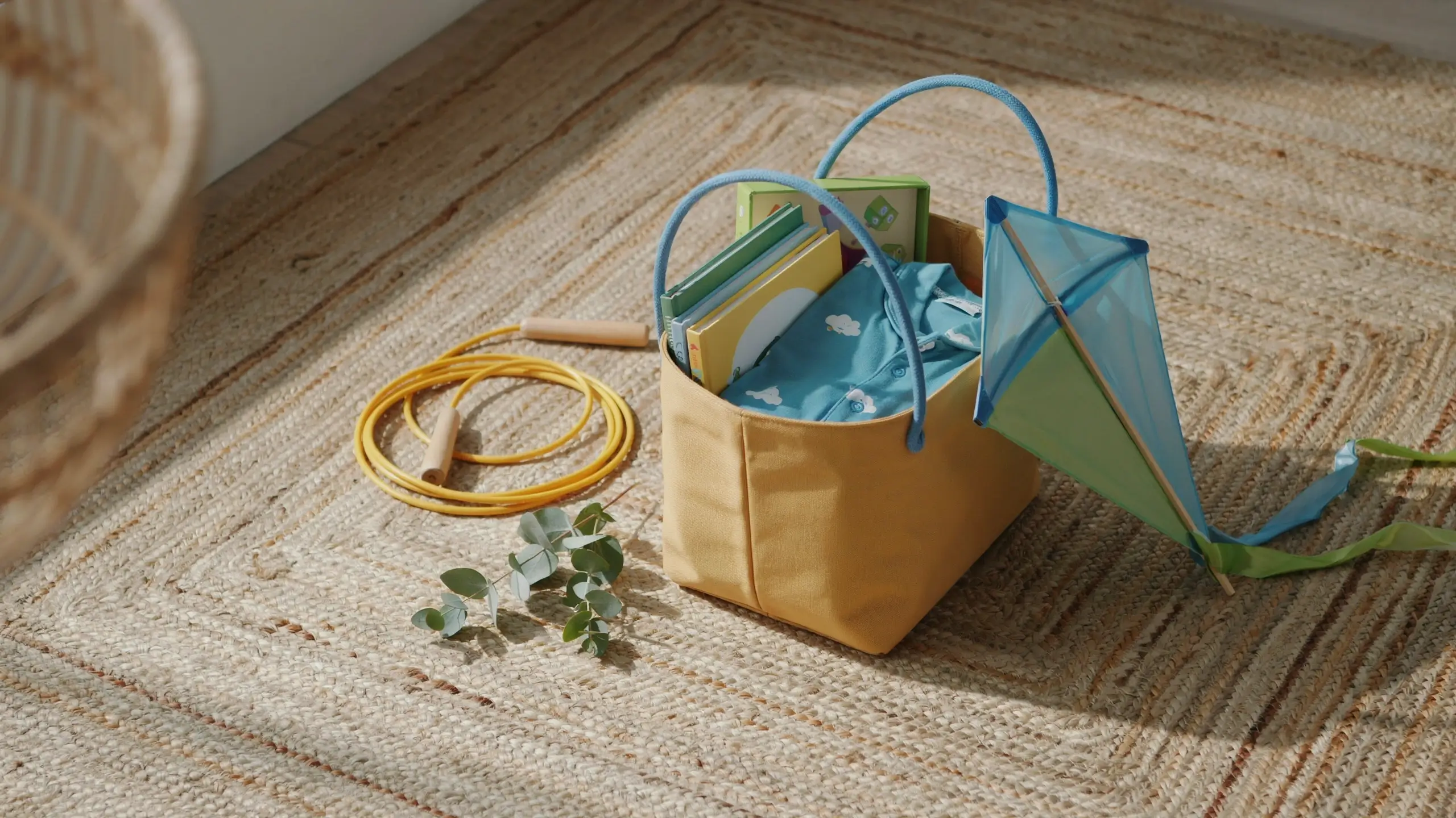 Non-candy Easter basket ideas featuring a yellow tote filled with children's books, a blue cloud-patterned shirt, and a kite, surrounded by a jump rope and eucalyptus on a woven rug.