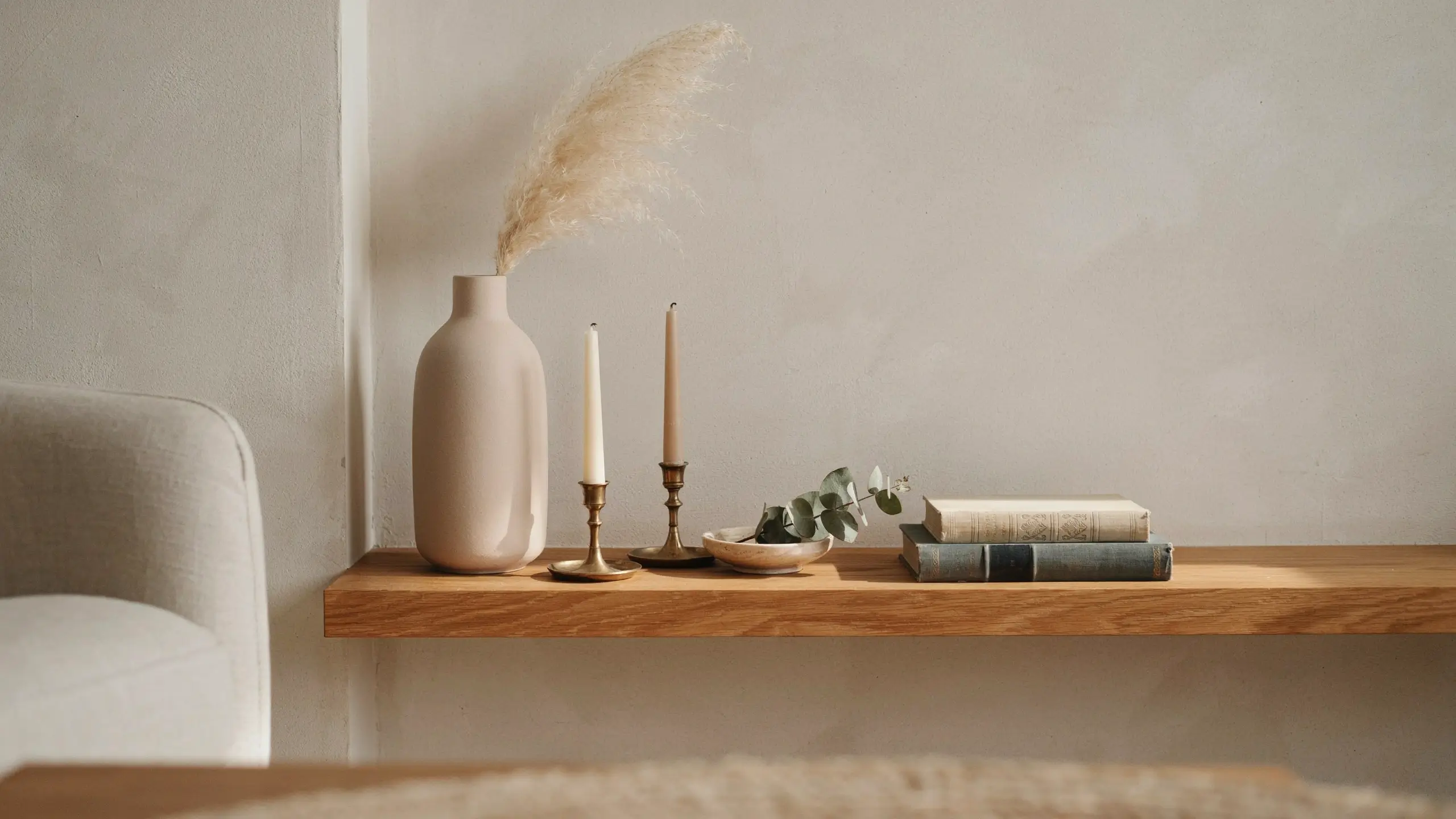 A minimalist floating wooden shelf styled with five decorative elements: a beige ceramic vase with dried pampas grass, two brass candle holders, a small bowl with eucalyptus leaves, and a stack of two vintage books, demonstrating the odd number styling rule for balanced interior home decor.