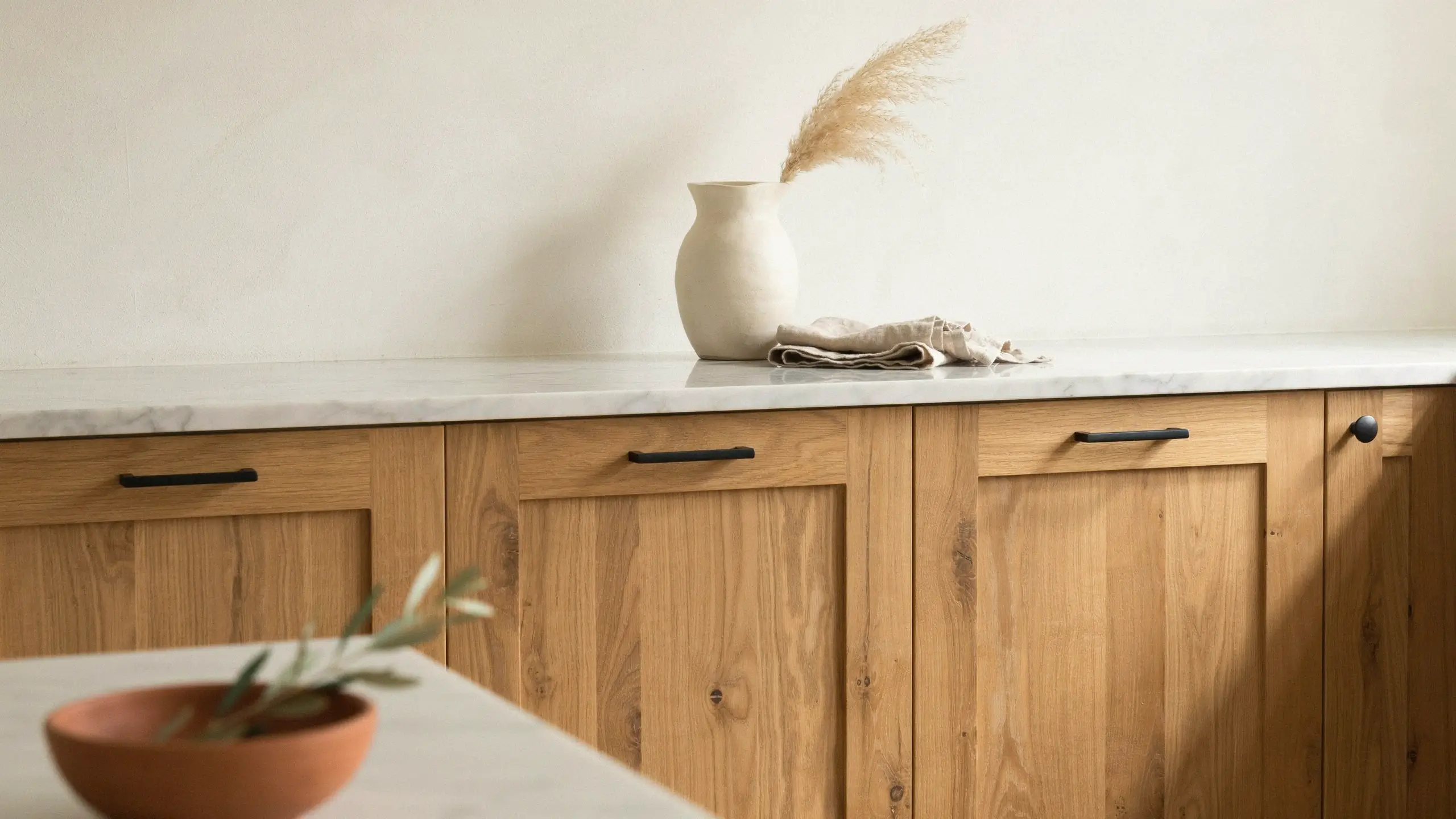 Light oak wood shaker cabinets with matte black drawer pulls, styled with dried pampas grass and a terracotta olive branch bowl, showcasing natural textures that beautifully complement a sage or olive green shaker front kitchen design.