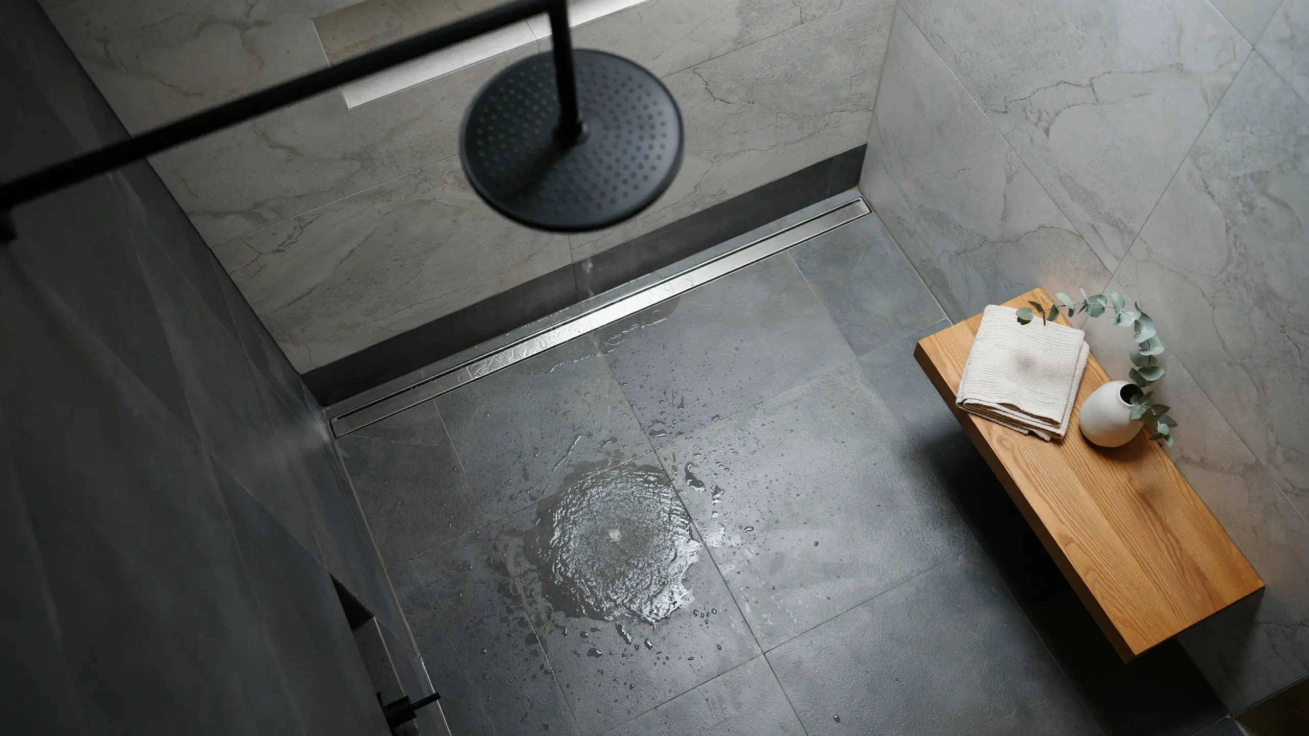 Modern minimalist wet room with black rain showerhead, grey stone floor tiles, linear drain, and wooden bench with eucalyptus.