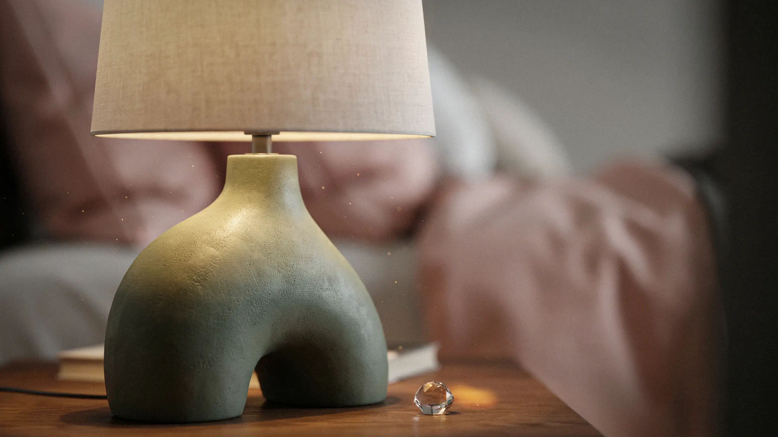 Close-up of a textured, organic-shaped sage green ceramic table lamp with a beige linen shade and a small clear crystal accent, sitting on a wooden nightstand in a cozy bedroom with soft pink blankets in the background.