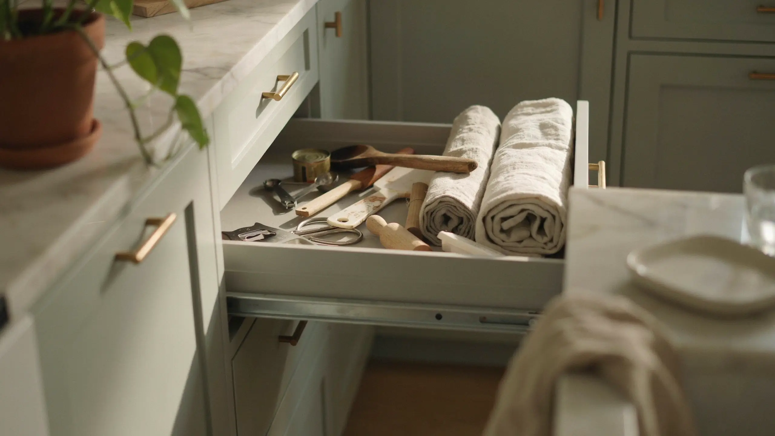 Open white kitchen drawer neatly arranged with wooden spoons, scissors, and rolled linen towels on a marble countertop.