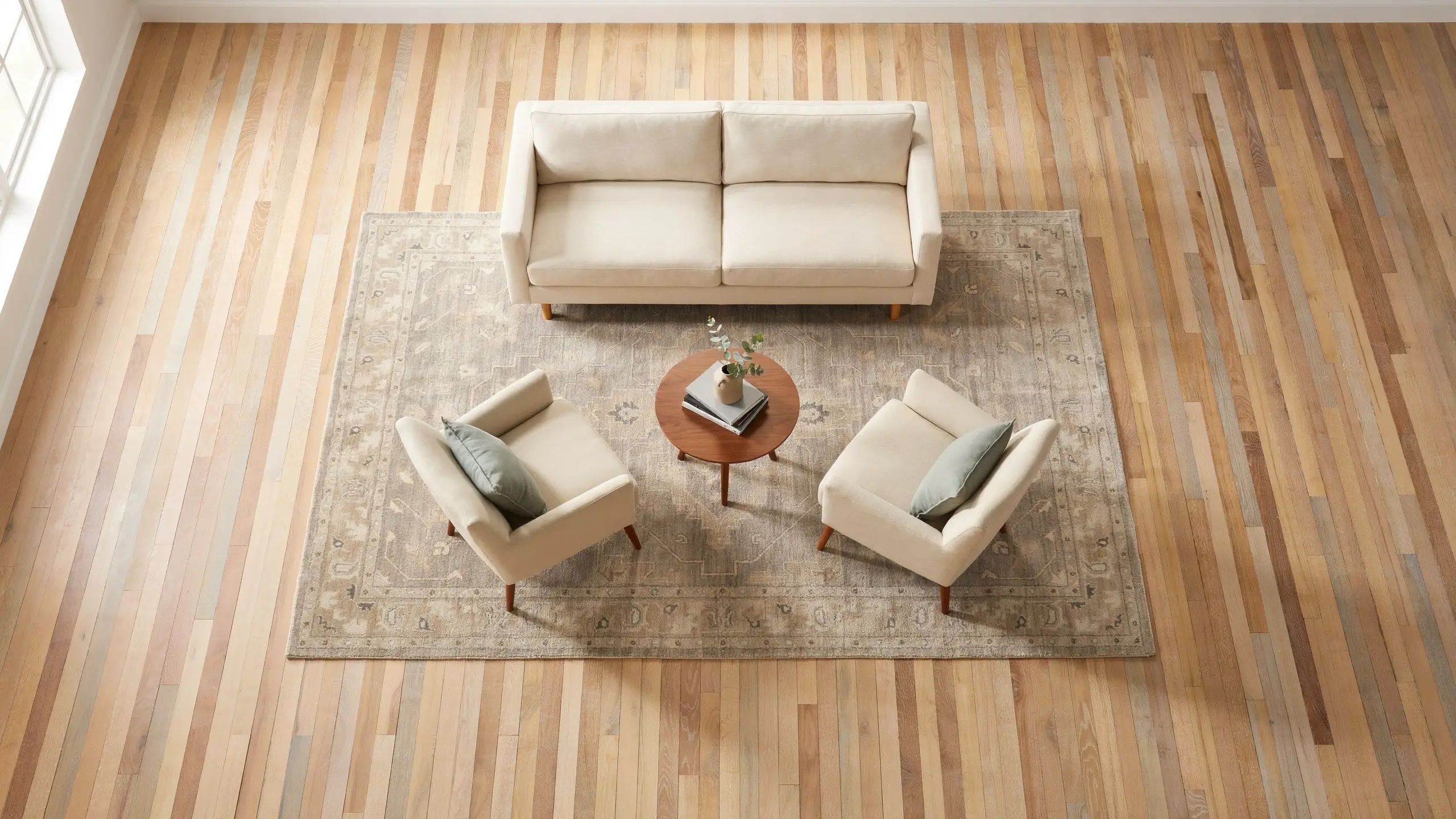 Top-down interior shot of a large area rug correctly positioned under a cream sofa, two matching accent armchairs, and a round wood coffee table to anchor a symmetrical living room layout.