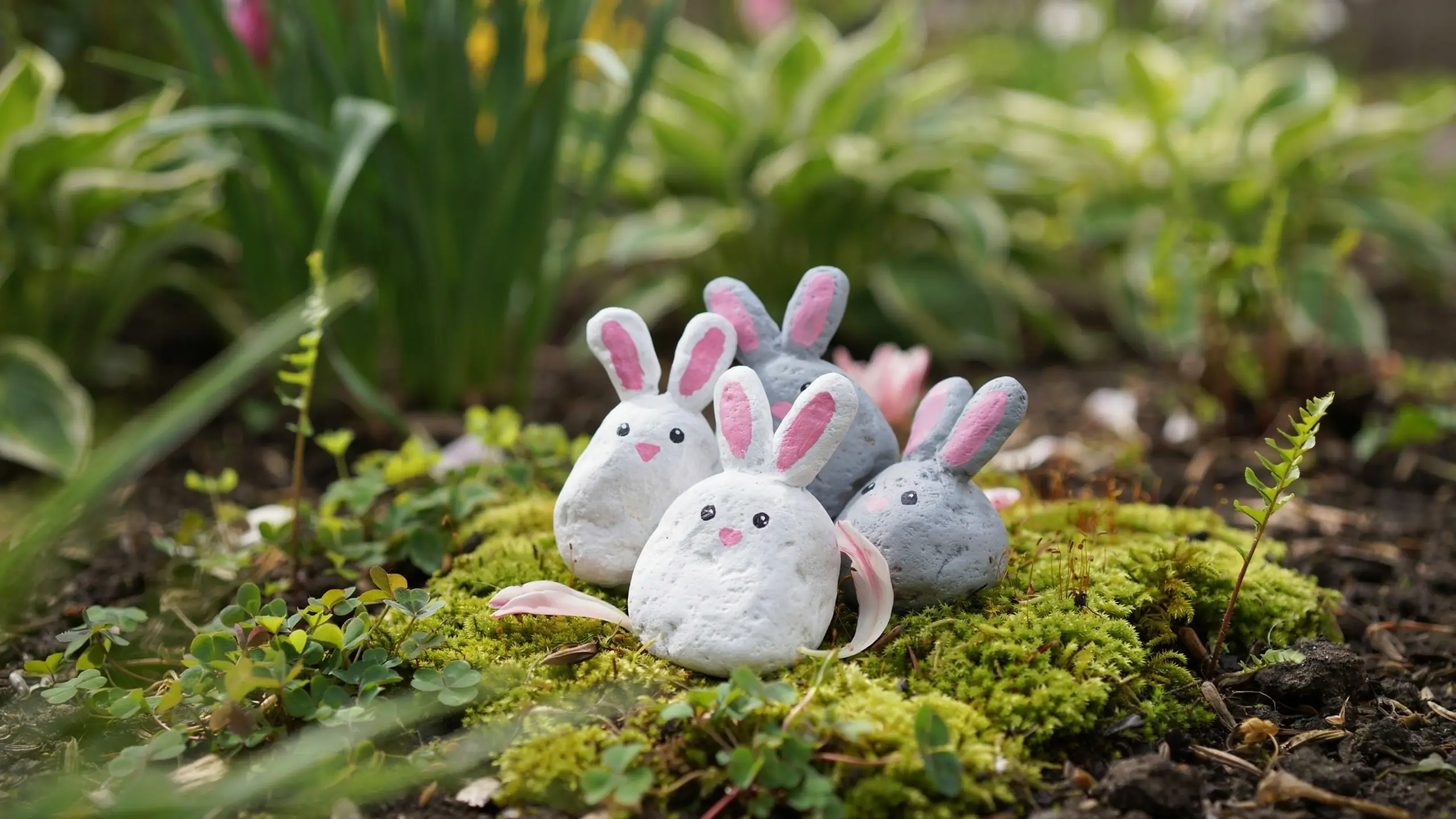 A charming cluster of four painted rock bunnies, featuring white and gray stones decorated with pink ears and tiny painted faces, nestled on vibrant green moss and clover in a spring garden bed.