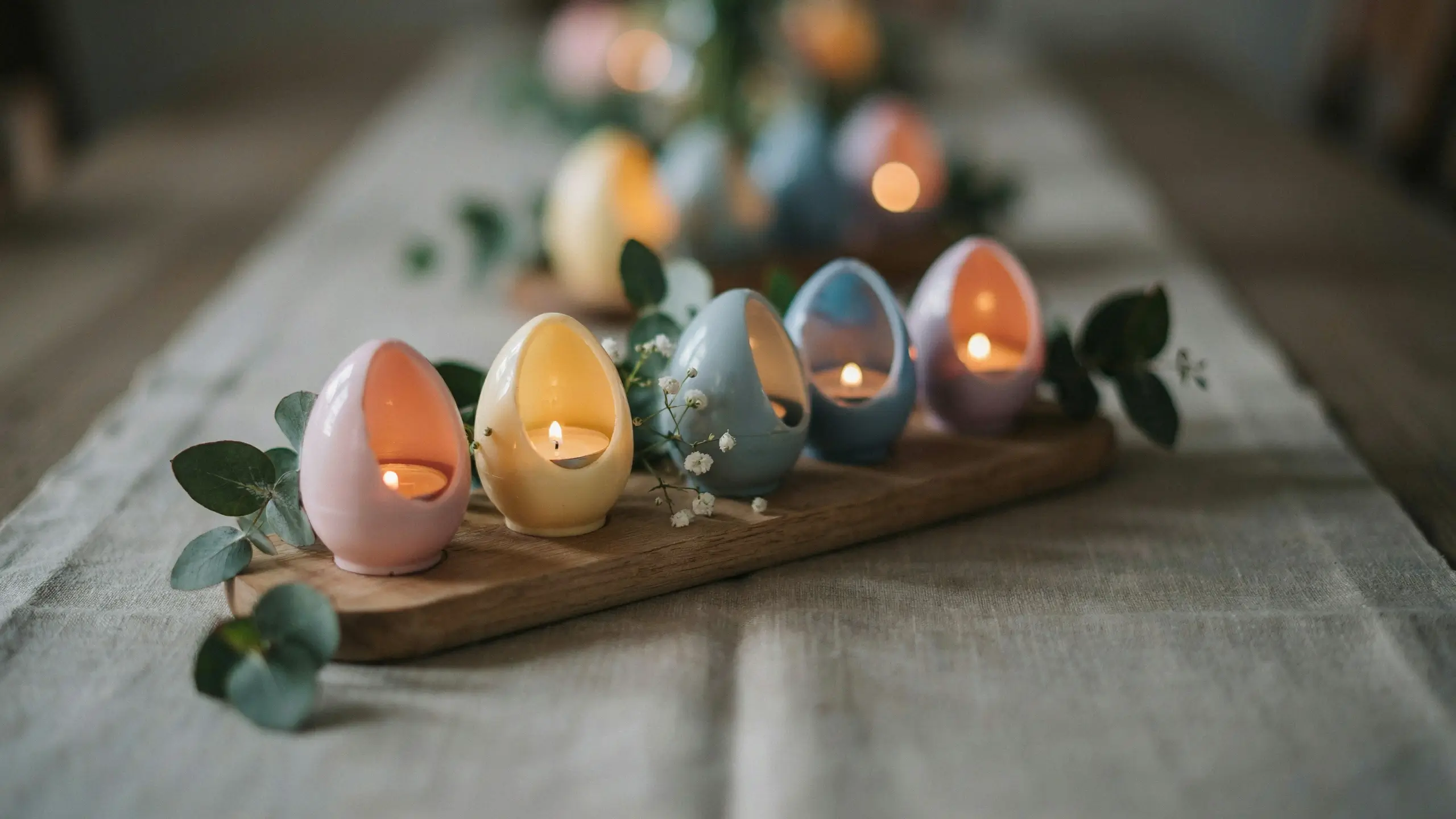 Five pastel-colored ceramic egg-shaped tea light holders with burning candles, arranged on a rustic wooden board and accented with fresh eucalyptus and baby's breath for a cozy Easter table centerpiece.