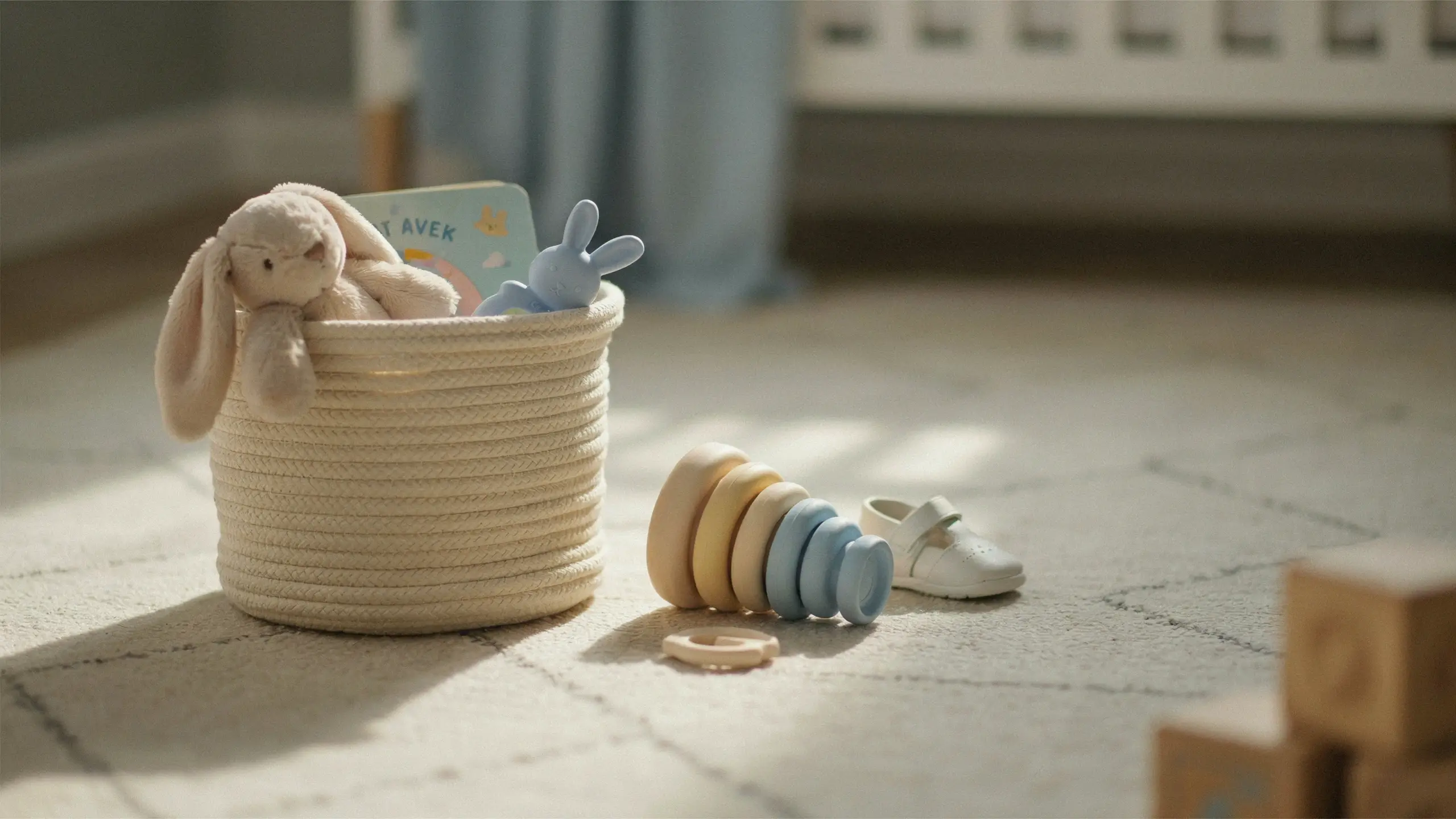 Pastel toy Easter basket for babies and toddlers featuring a plush bunny, blue silicone rattle, board book, and wooden stacking rings scattered on a cozy nursery rug.