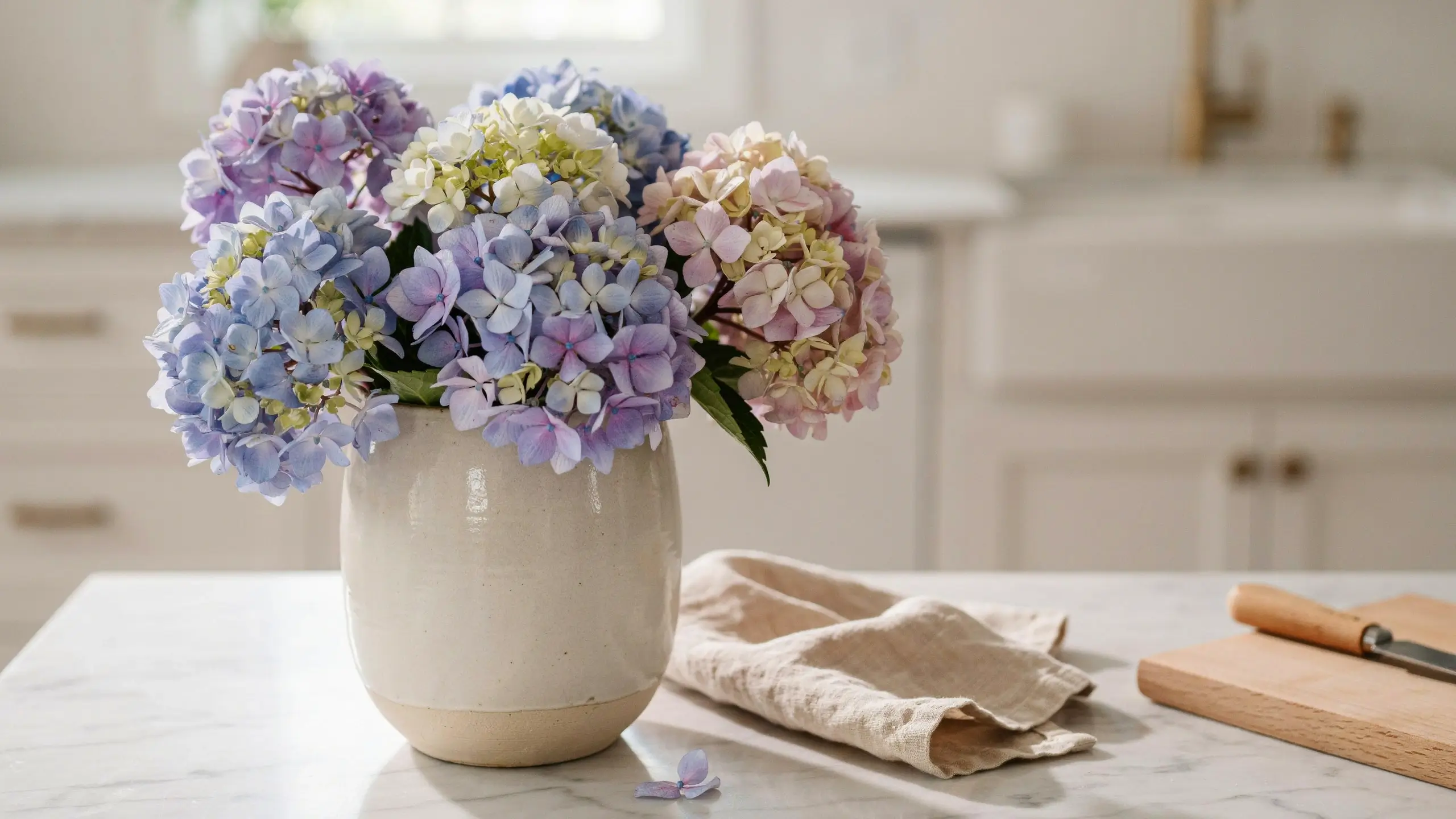 Hydrangea stems in pastel blue, purple, and pink tones arranged in a cream ceramic vase on a white marble kitchen counter beside a wooden cutting board.