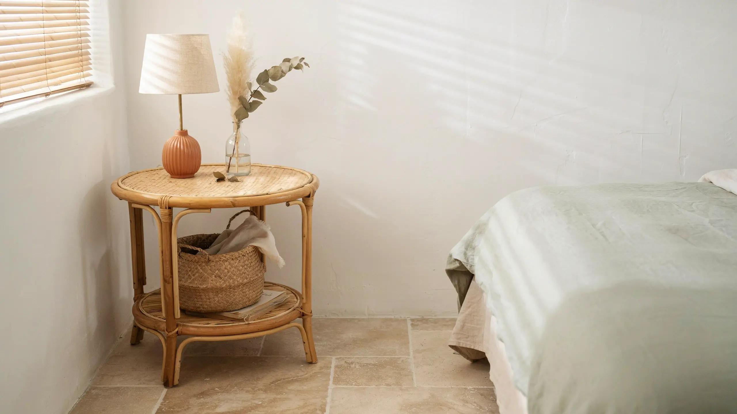 Round rattan cane side table in a sunlit bedroom, styled with a terracotta lamp base, a glass vase with pampas grass, and a woven storage basket on the lower shelf. 