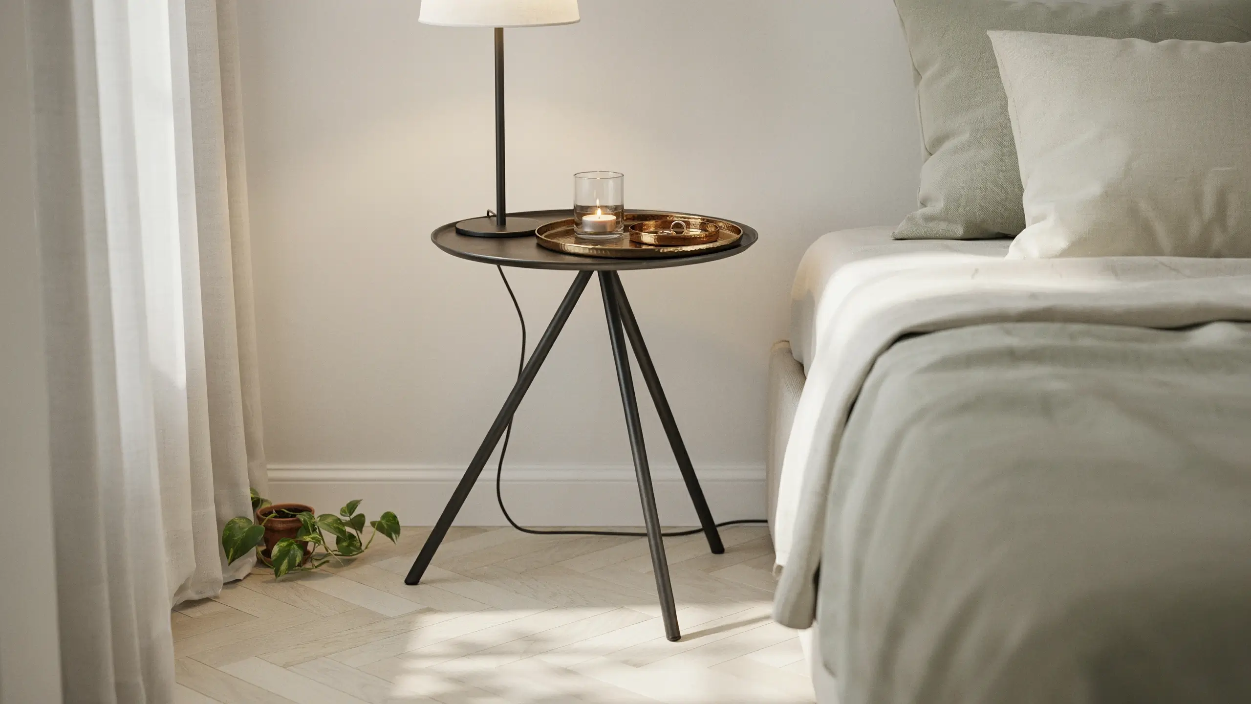 A black tripod plant stand creatively repurposed as a minimalist bedside table, holding a lit candle, a small gold tray, and a table lamp, positioned next to a bed with neutral grey pillows in a bright bedroom.